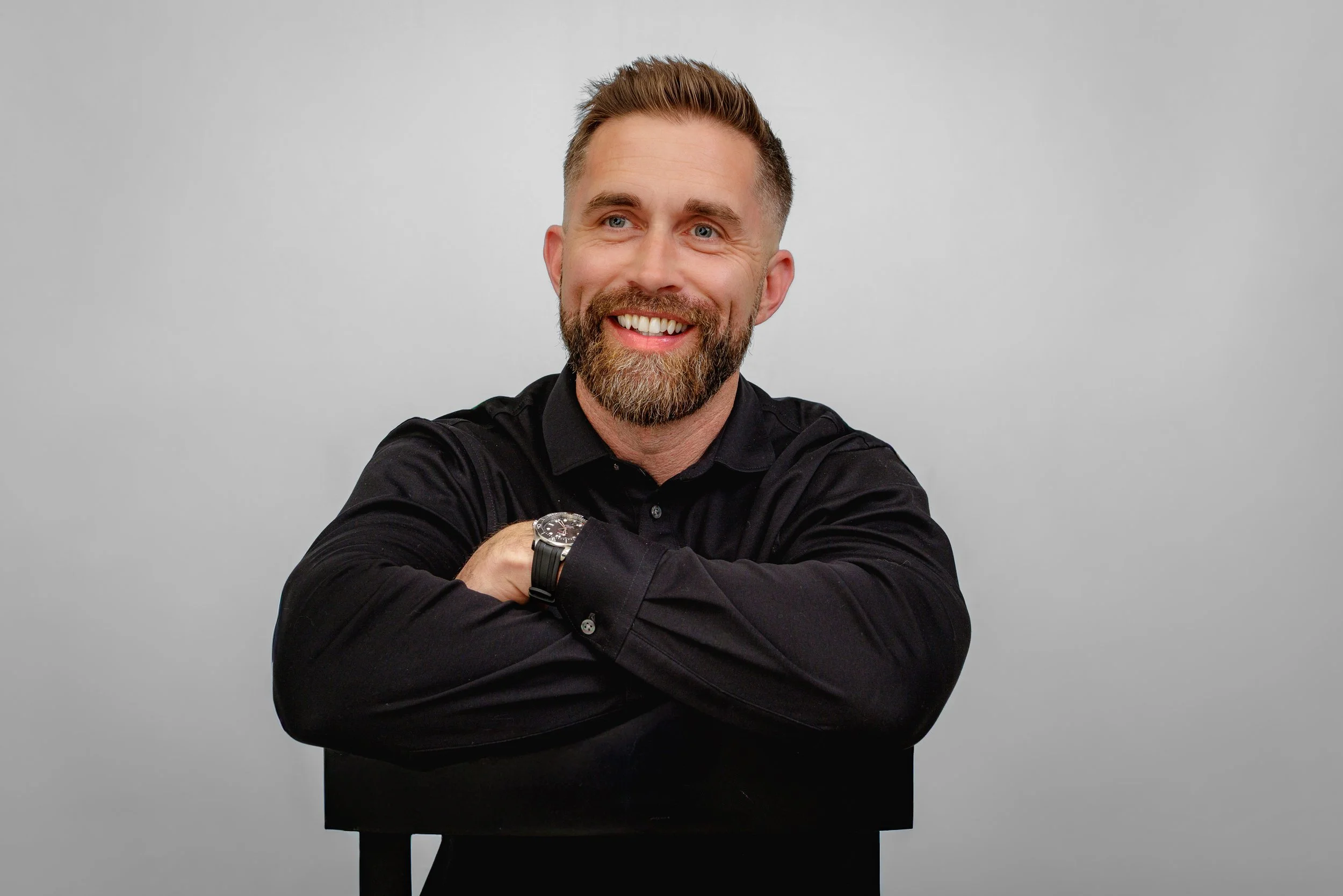 A smiling man with a beard and short hair, wearing a black shirt, sitting with crossed arms against a plain light gray background.