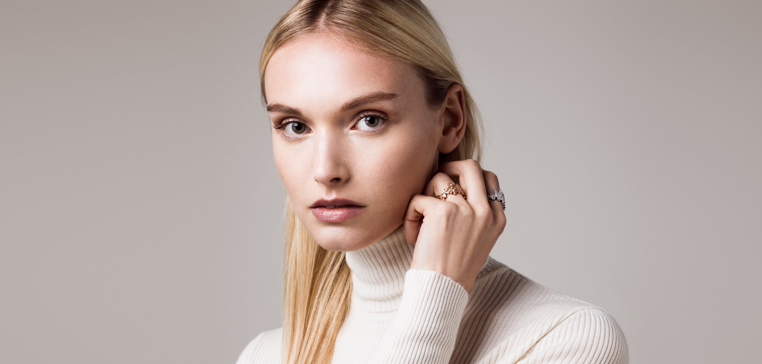 Nahaufnahme eines jungen Mädchens mit blonden Haaren, weißem Rollkragenpullover, trägt verschiedene Ringe an der Hand und schaut direkt in die Kamera, neutraler Hintergrund