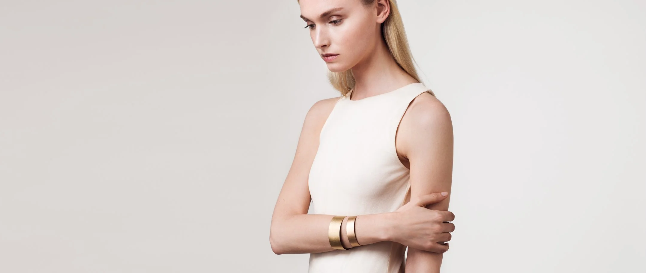 Eine junge Frau mit blonden Haaren trägt ein weißes ärmelloses Kleid und mehrere goldene Armbänder, steht vor einem neutralen, hellgrauen Hintergrund.