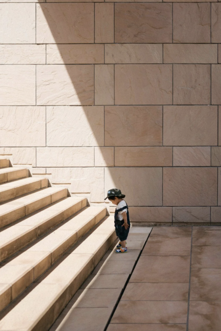 A young boy standing at the bottom of staircase steps made of beige stone, looking down, with a large stone wall in the background partially covered in shadow.