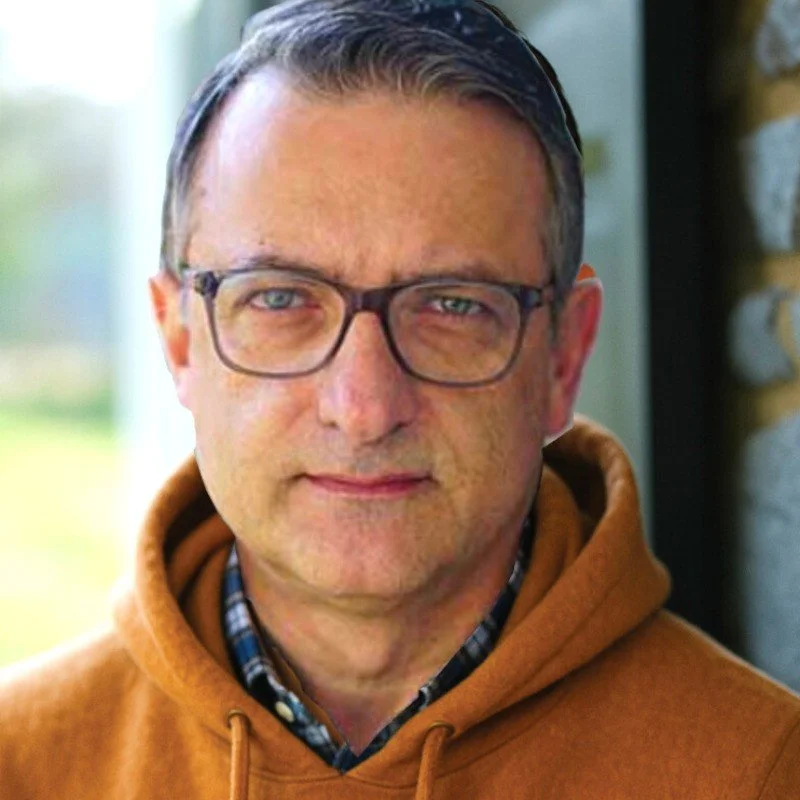 A man wearing glasses and a brown hoodie, standing outdoors with a blurred background.