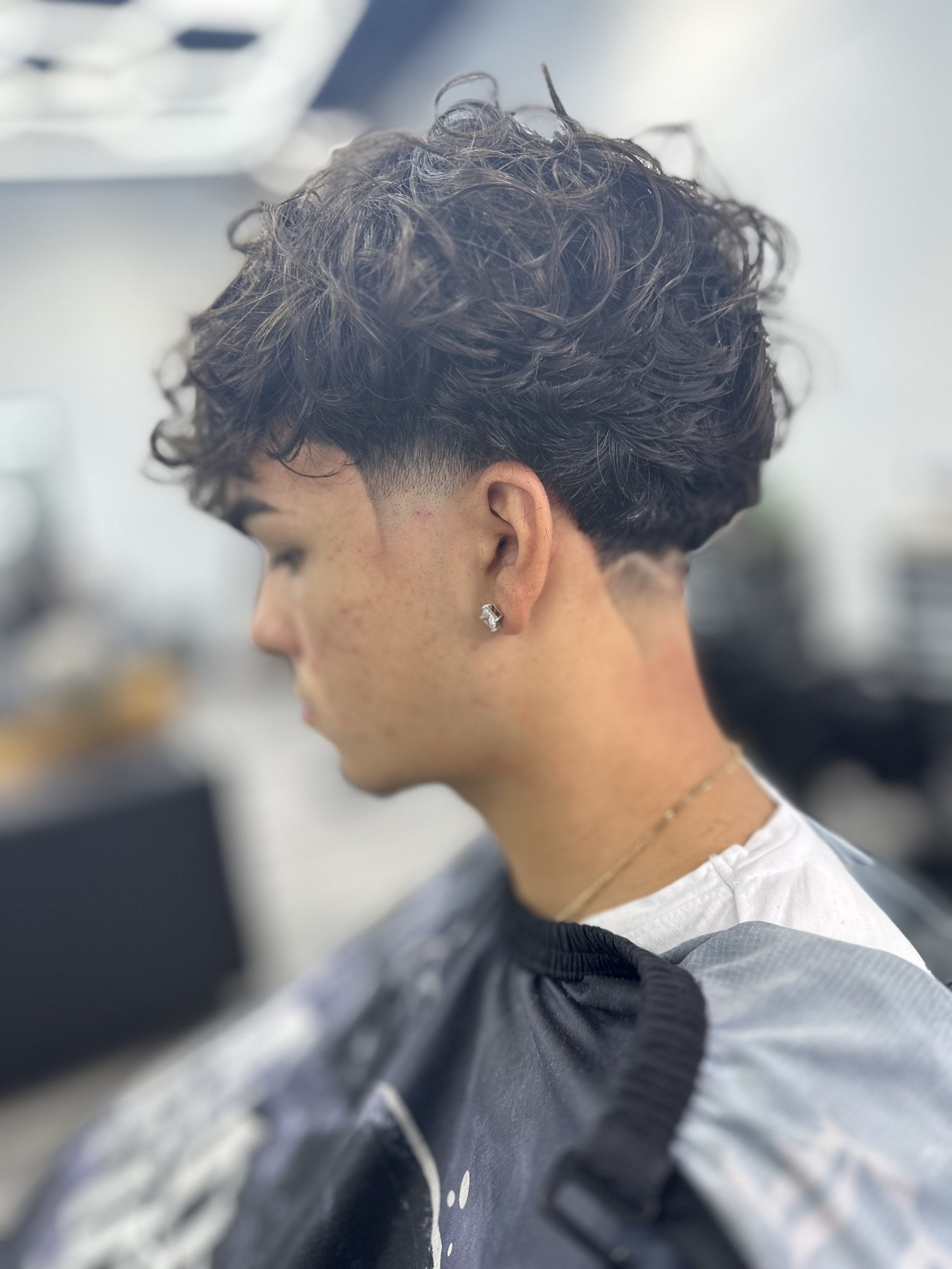 Person with curly hairstyle and undercut, wearing a white shirt and an earring, in a modern indoor setting.