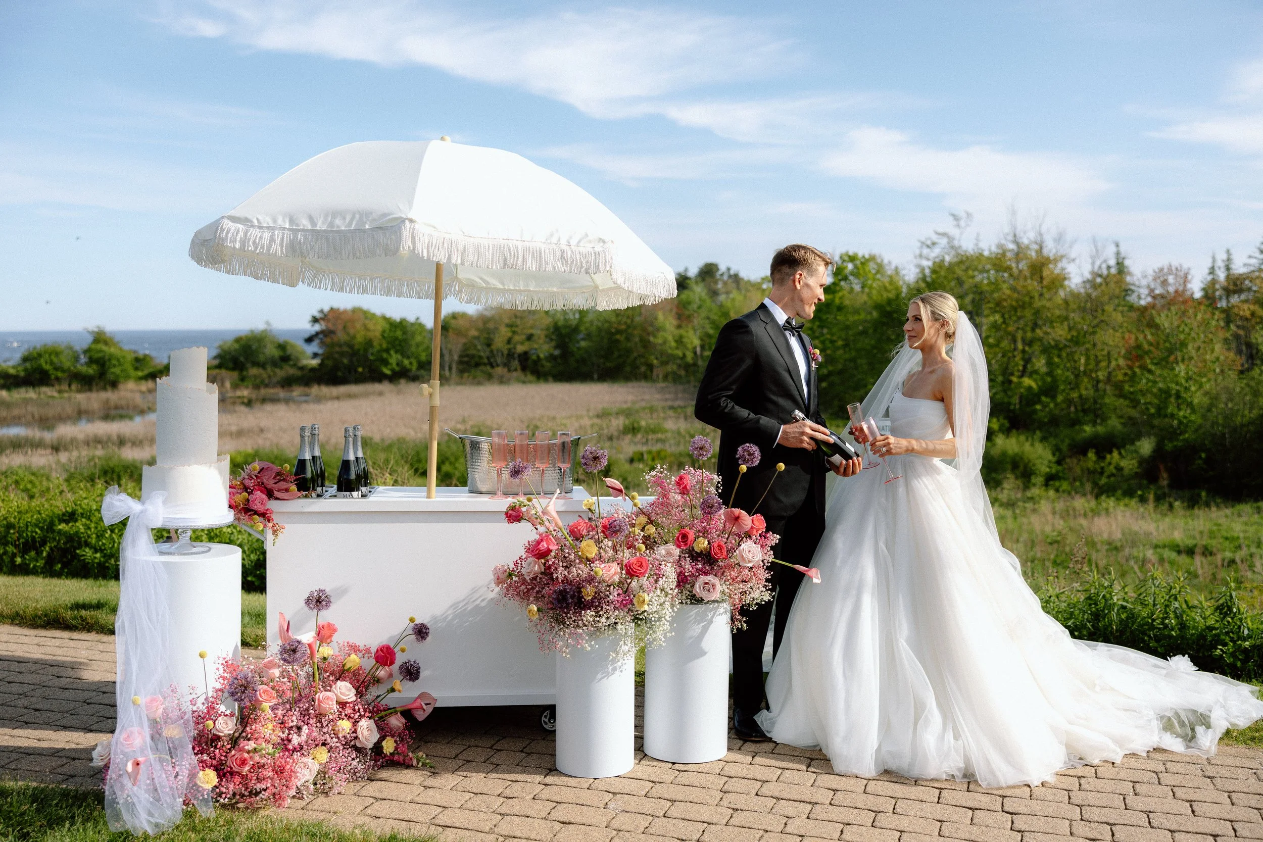 Bride and groom at outdoor wedding reception with a table of drinks, flowers, and a large white umbrella, set against a green landscape and blue sky.