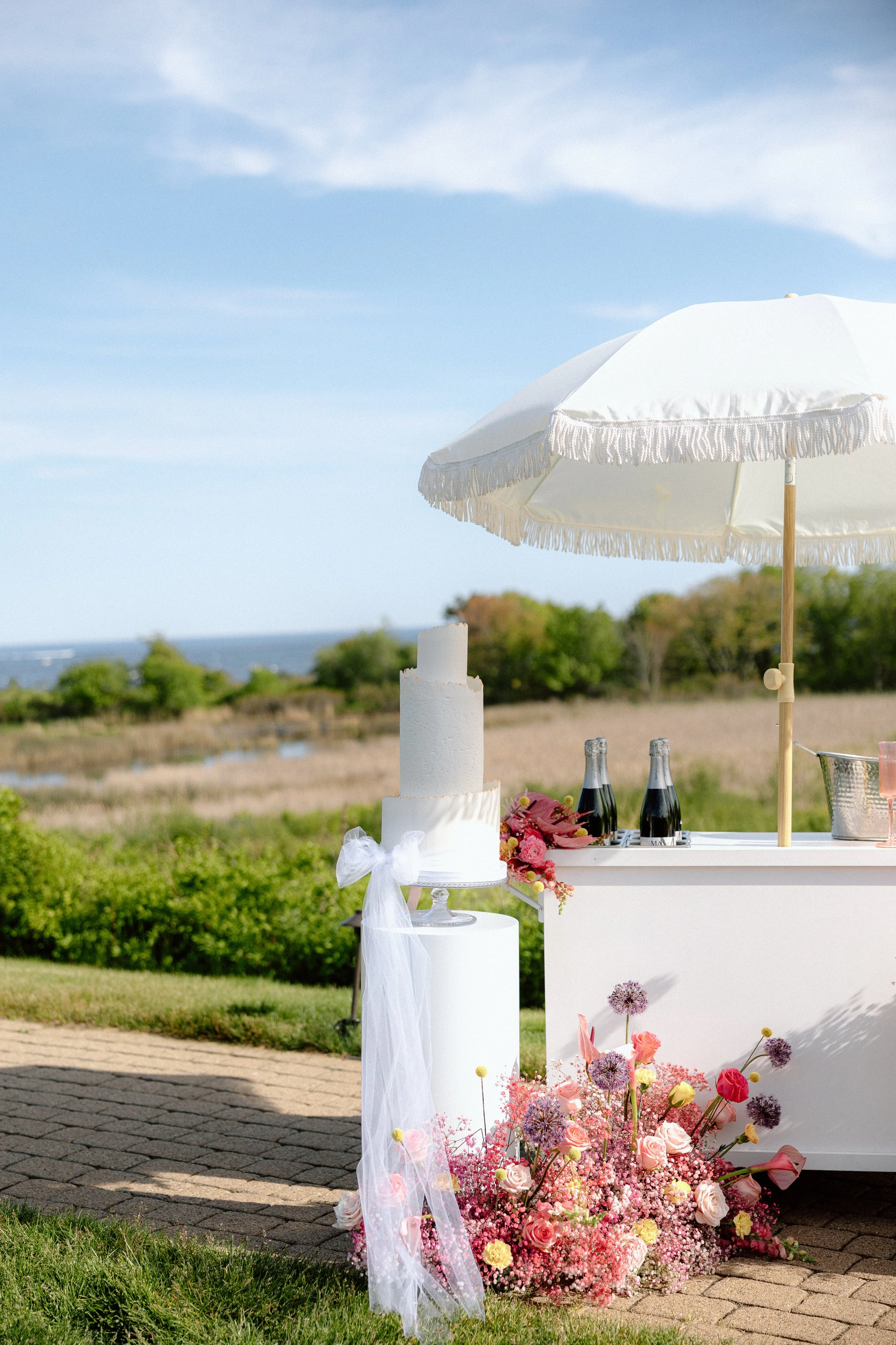 A wedding cake decorated with white icing and a white bow, surrounded by pink flowers, set on a white platform outdoors. There's a large white parasol shading the area and a table with bottles of champagne in the background, overlooking a scenic landscape with fields and trees on a clear day.