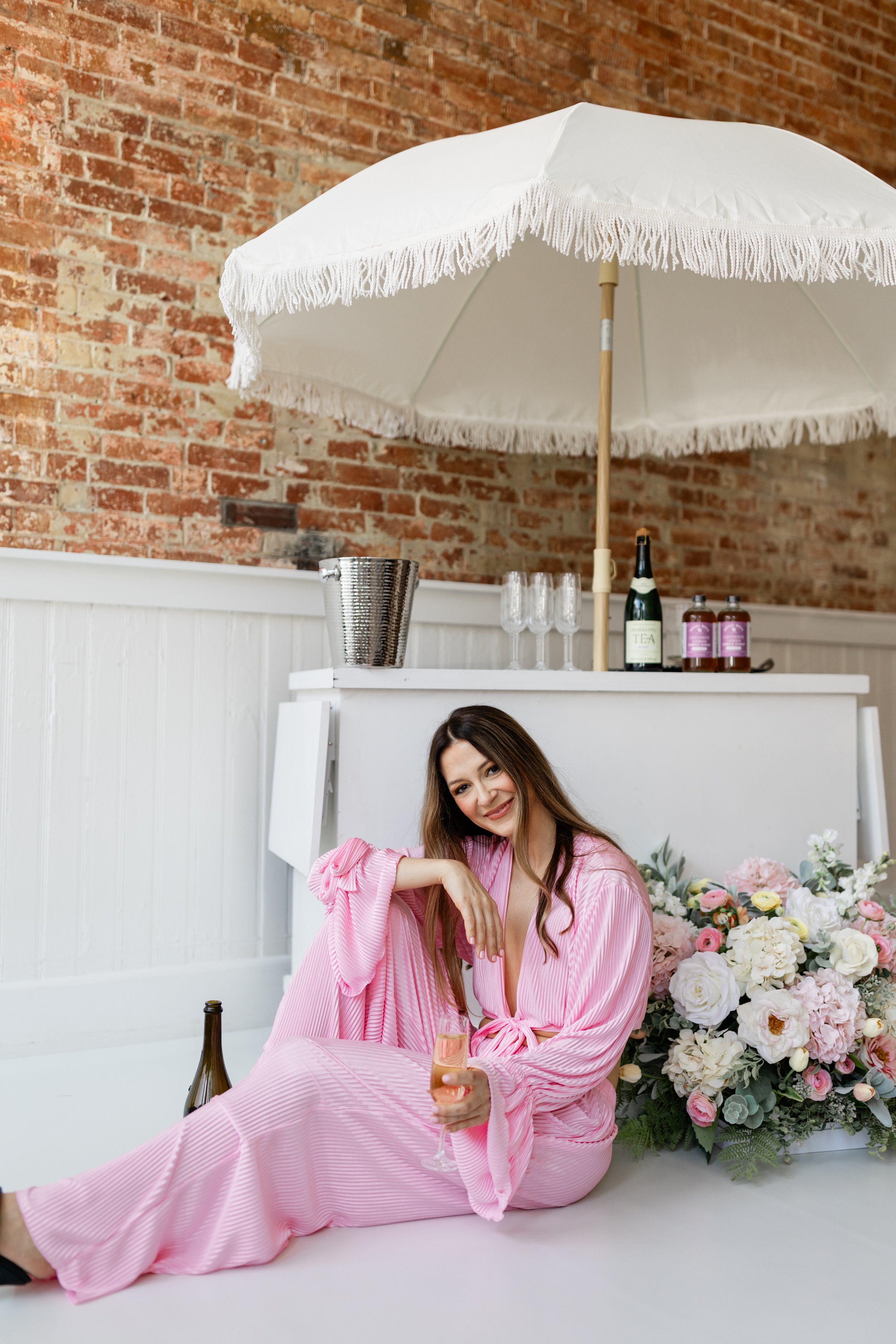 Rochelle (owner) in pink matching set sitting on the floor, holding a glass of rosé wine, with a bottle nearby, in a setting with a large white umbrella, a bouquet of pink and white flowers, and a brick wall in the background.