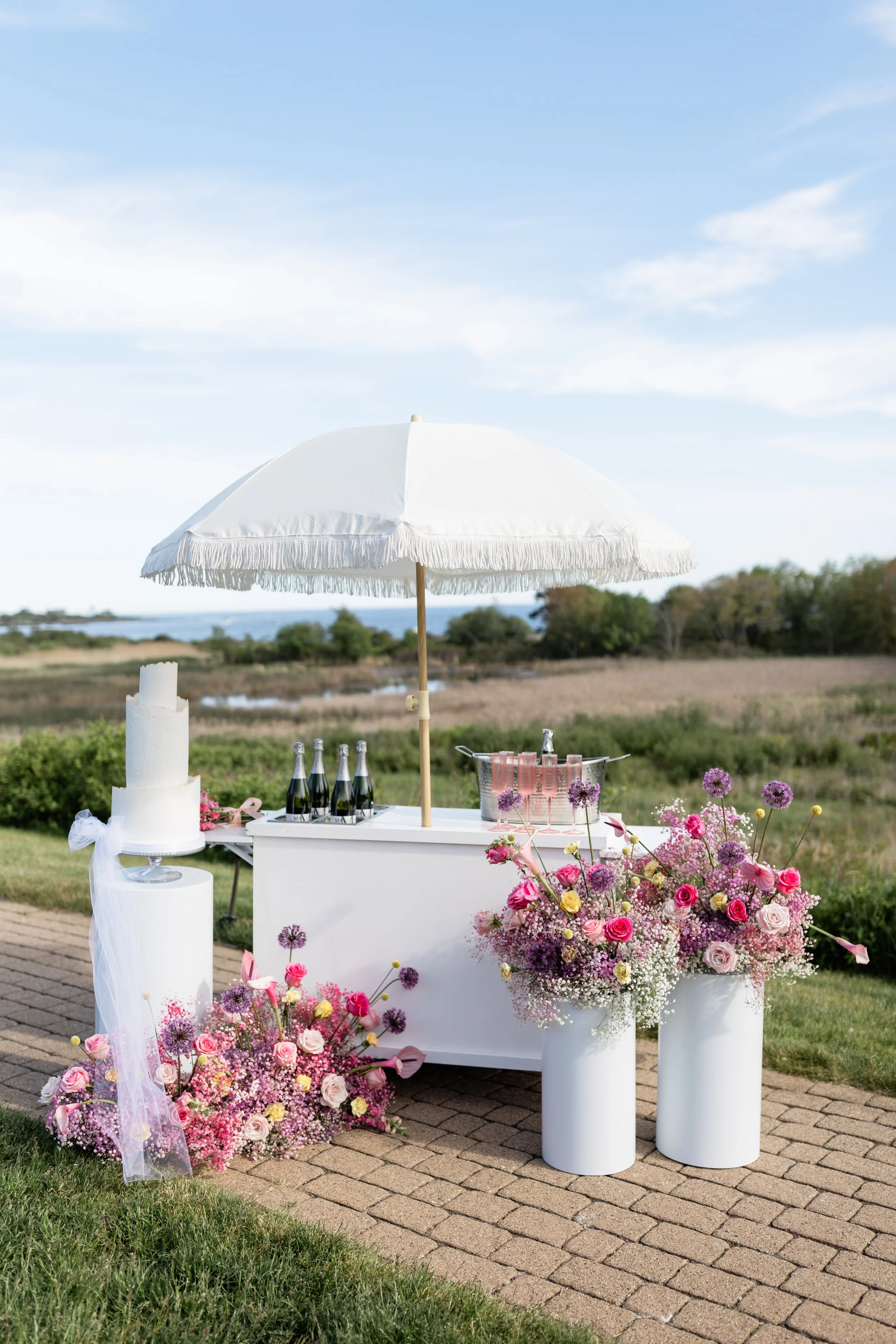 A flower-filled outdoor bar setup with a white umbrella, champagne bottles, a white cake, and floral arrangements on white pedestals and a white table, set against a grassy landscape with trees and a river in the background.