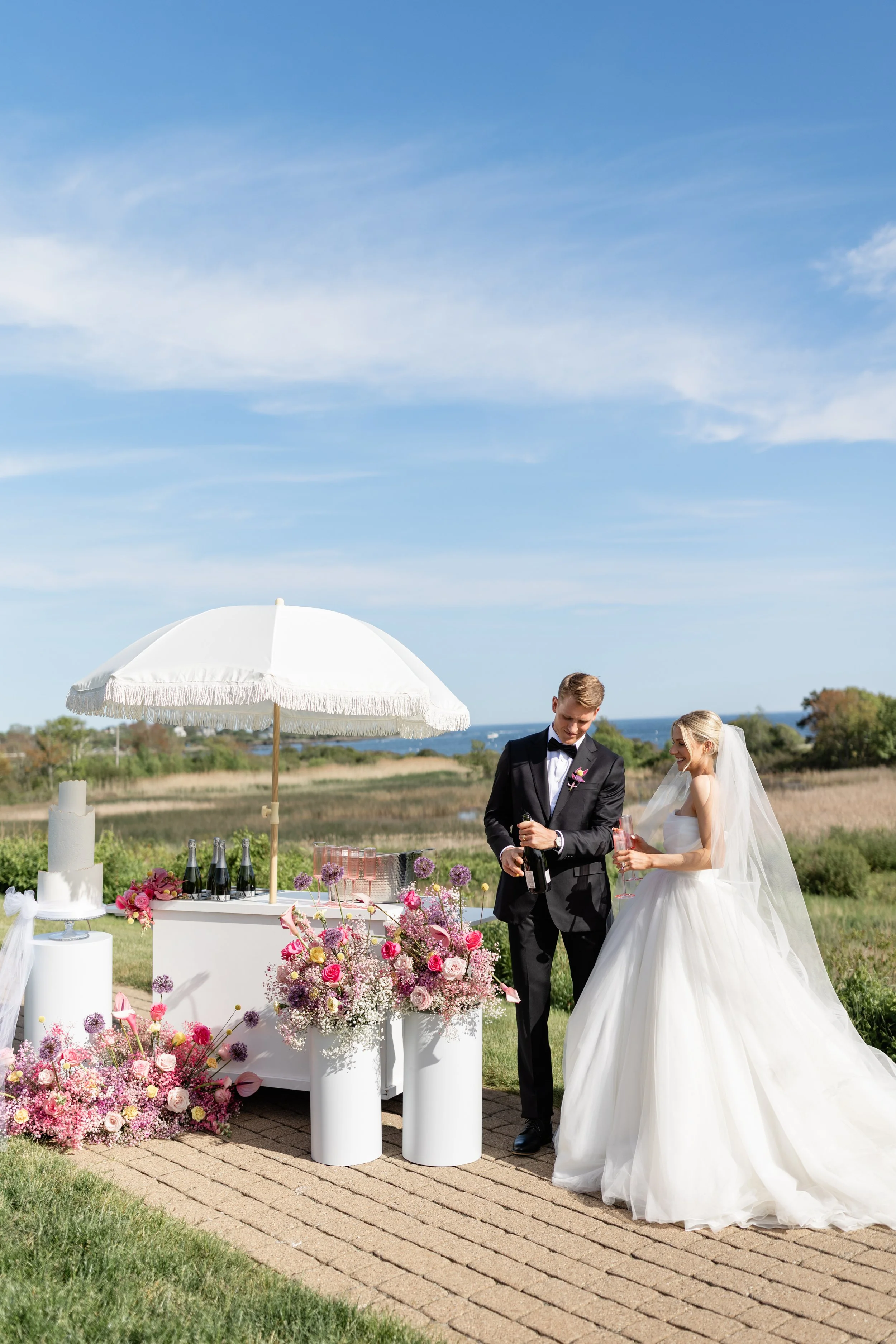 A bride and groom in wedding attire stand outdoors next to a white bar with pink and purple floral decorations, champagne bottles, and glasses, under a white parasol on a sunny day with a grassy landscape and blue sky in the background.