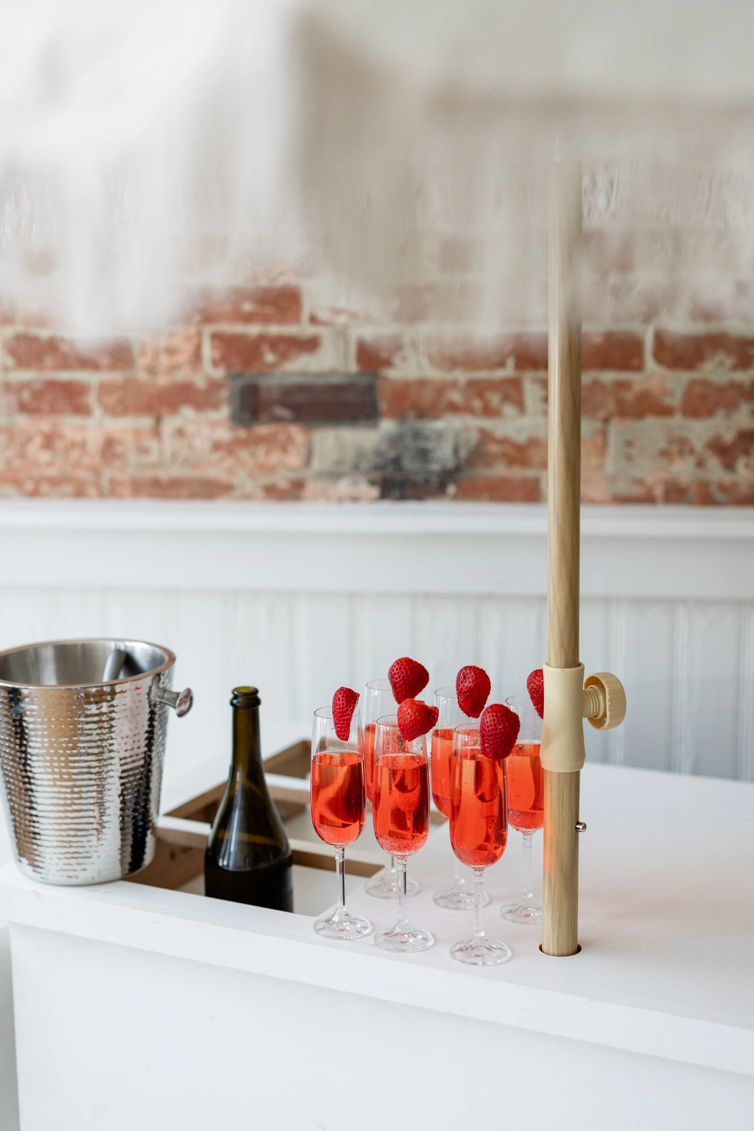 Six glasses of pink champagne with strawberries on top, a champagne bottle, and an ice bucket on a white bar or table against a brick wall.