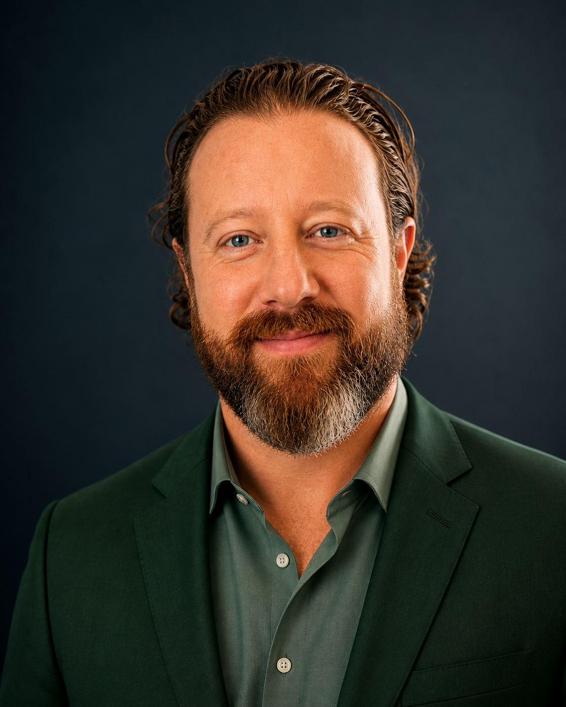 Headshot of a middle-aged man with light skin, curly brown hair, blue eyes, and a full beard, wearing a dark green suit and matching shirt, smiling against a dark background.