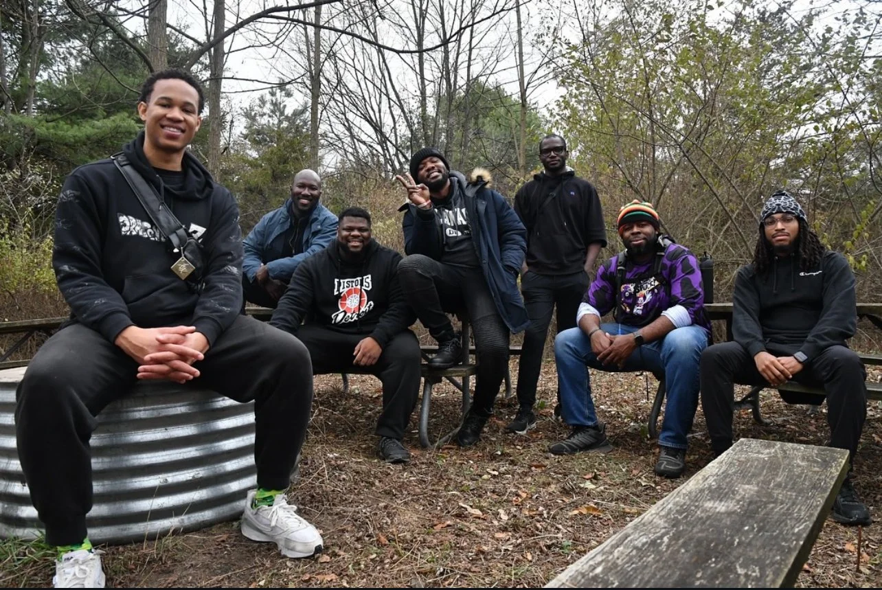 Seven people smiling and posing together at a rest stop on a wooded trail, seated on benches and a metal drum, surrounded by bare trees and fallen leaves