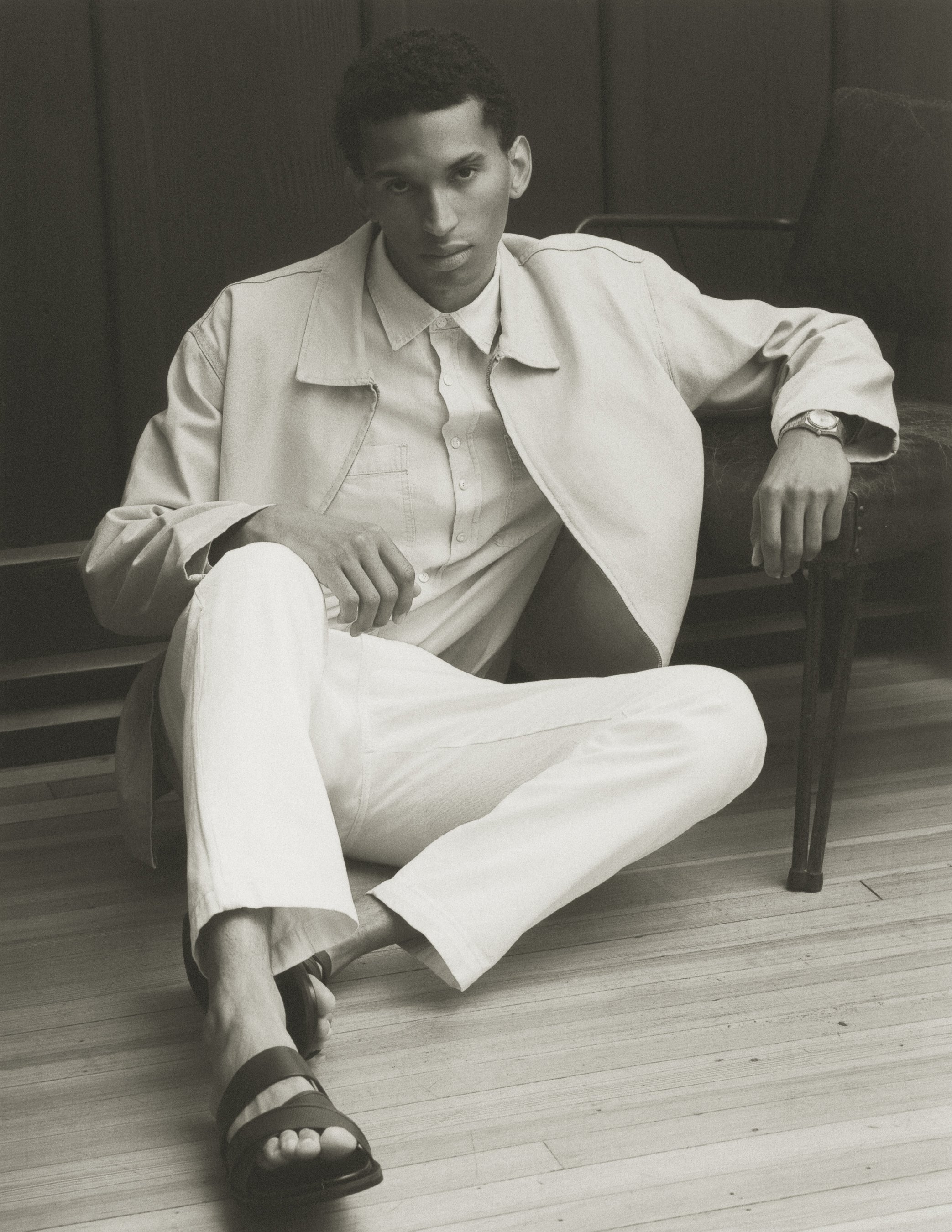 A young man with short dark hair, wearing a light-colored denim jacket, white shirt, white pants, and black sandals, sitting on a chair with a wooden floor and dark wall behind him.