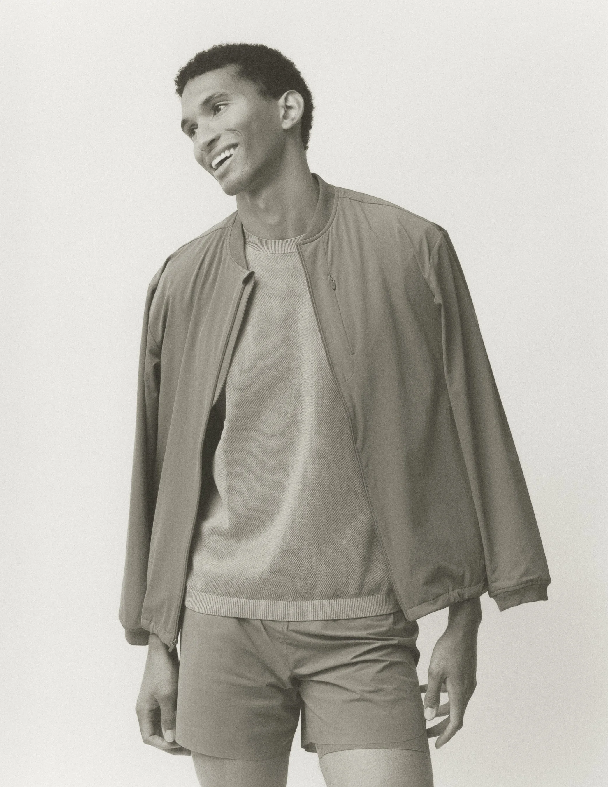 A young man smiling, wearing a casual jacket over a t-shirt, standing against a plain wall.