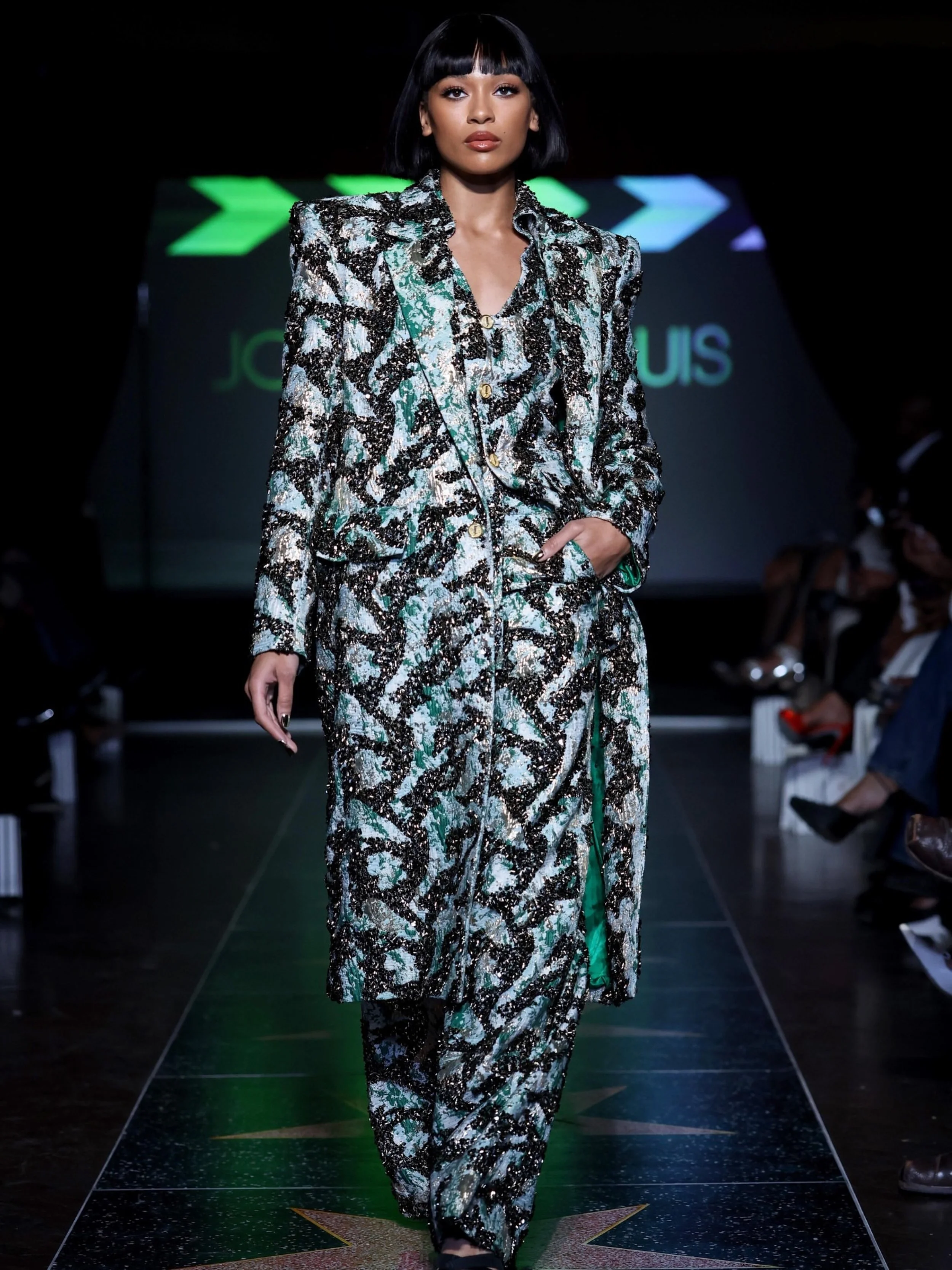 A female model with straight black hair and bangs walks down a runway wearing a matching outfit with a black, white, green, and blue abstract pattern. The background features a runway and audience, with a screen displaying the Louis Vuitton logo.