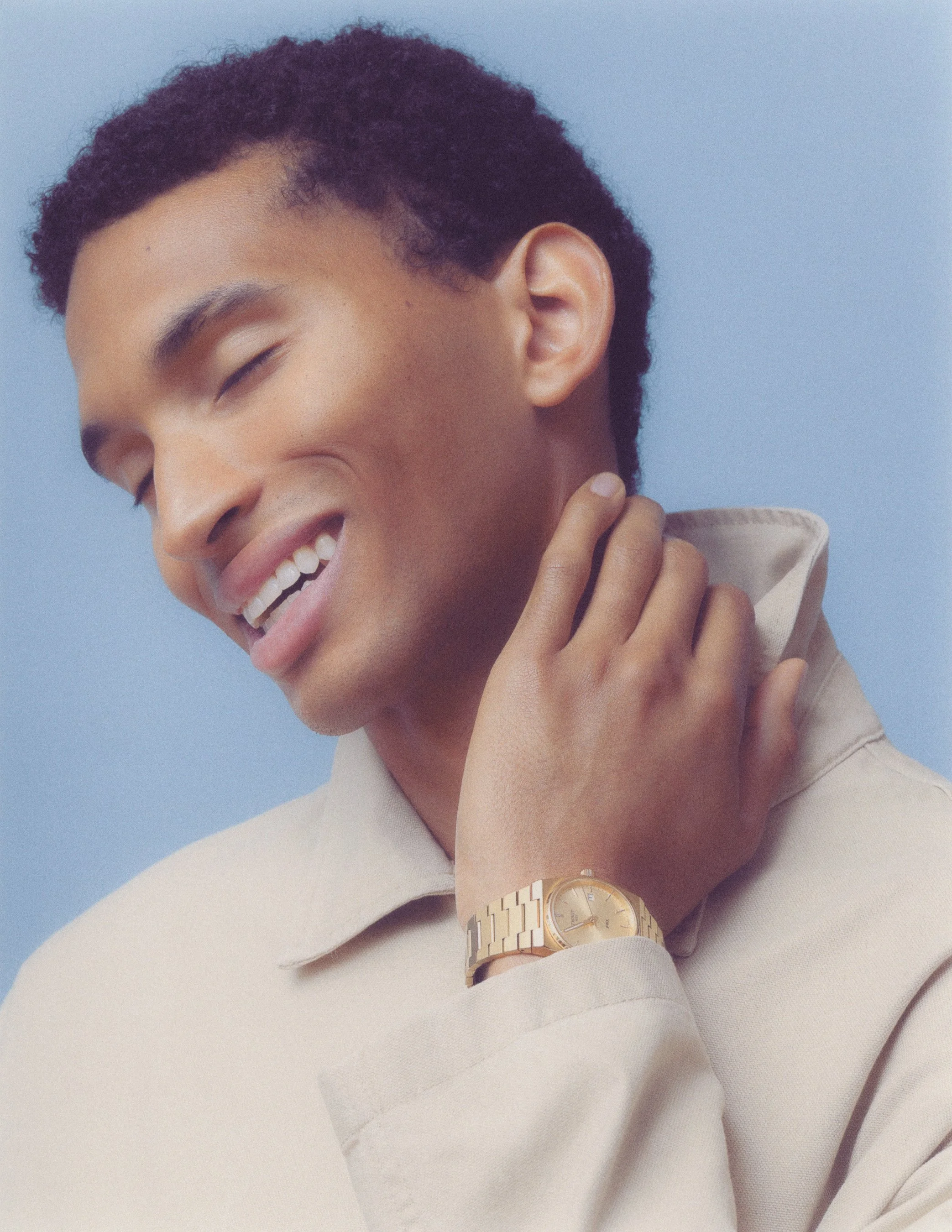 A smiling person with curly short hair, wearing a beige shirt, a wristwatch, and touching their neck. The background is a plain light blue.