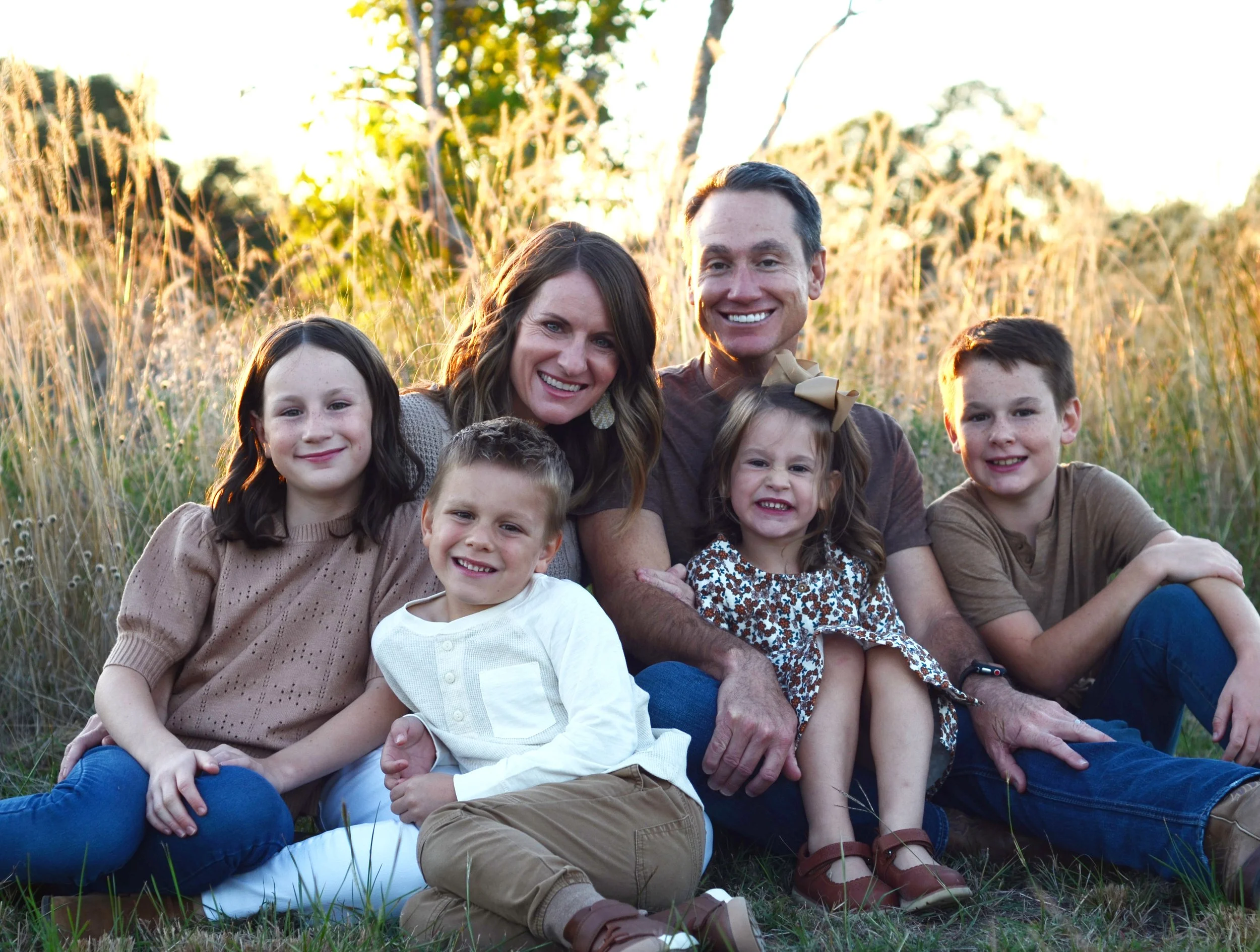 A smiling family of seven sitting outdoors in a grassy field during sunset, posing for a group photo.