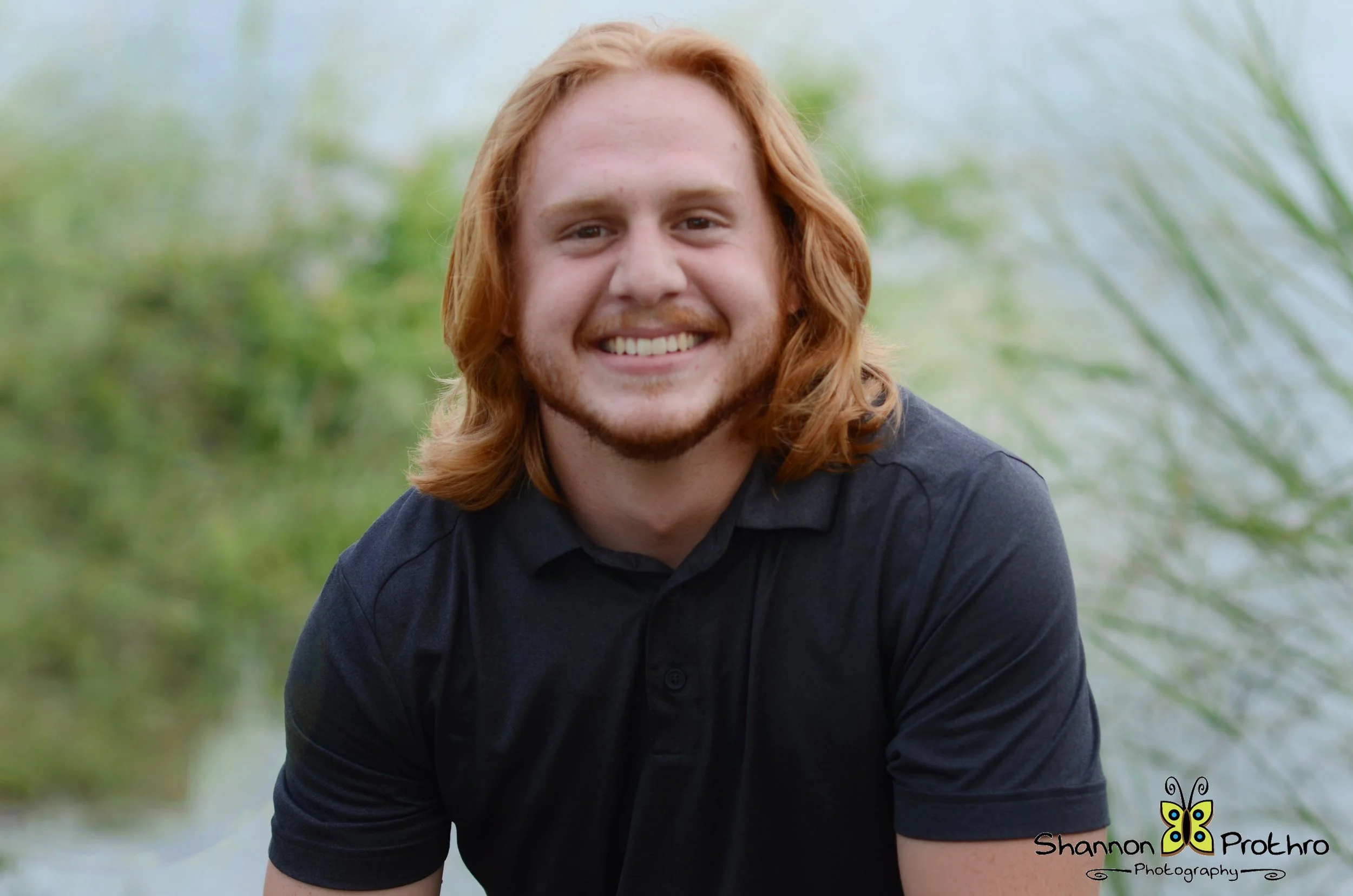 Young man with long red hair and a beard smiling outdoors with green foliage in the background.