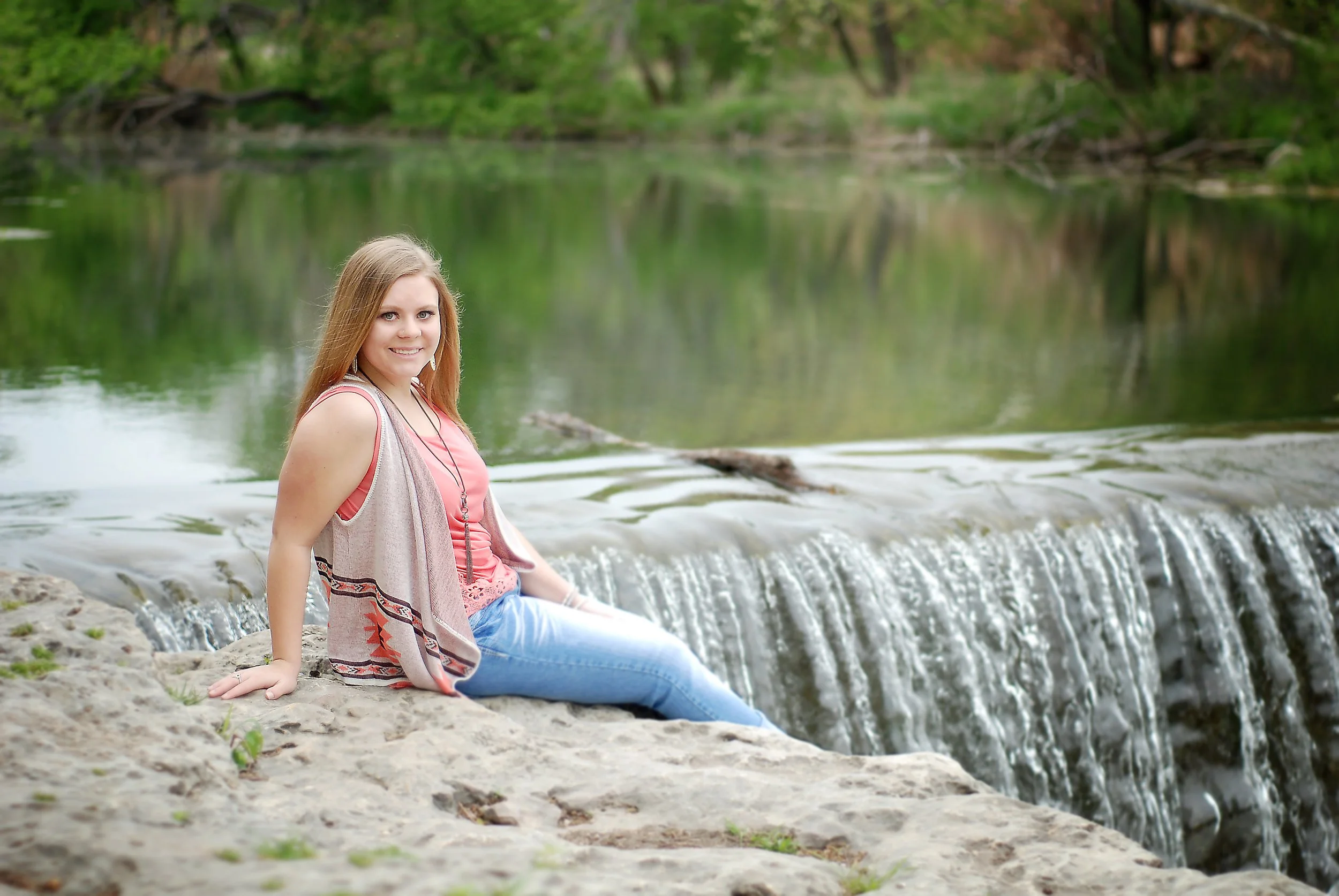 A young woman with long hair, wearing a pink top, light vest, and blue jeans, sitting on a rock near a small waterfall in a natural setting with trees reflected in the water.