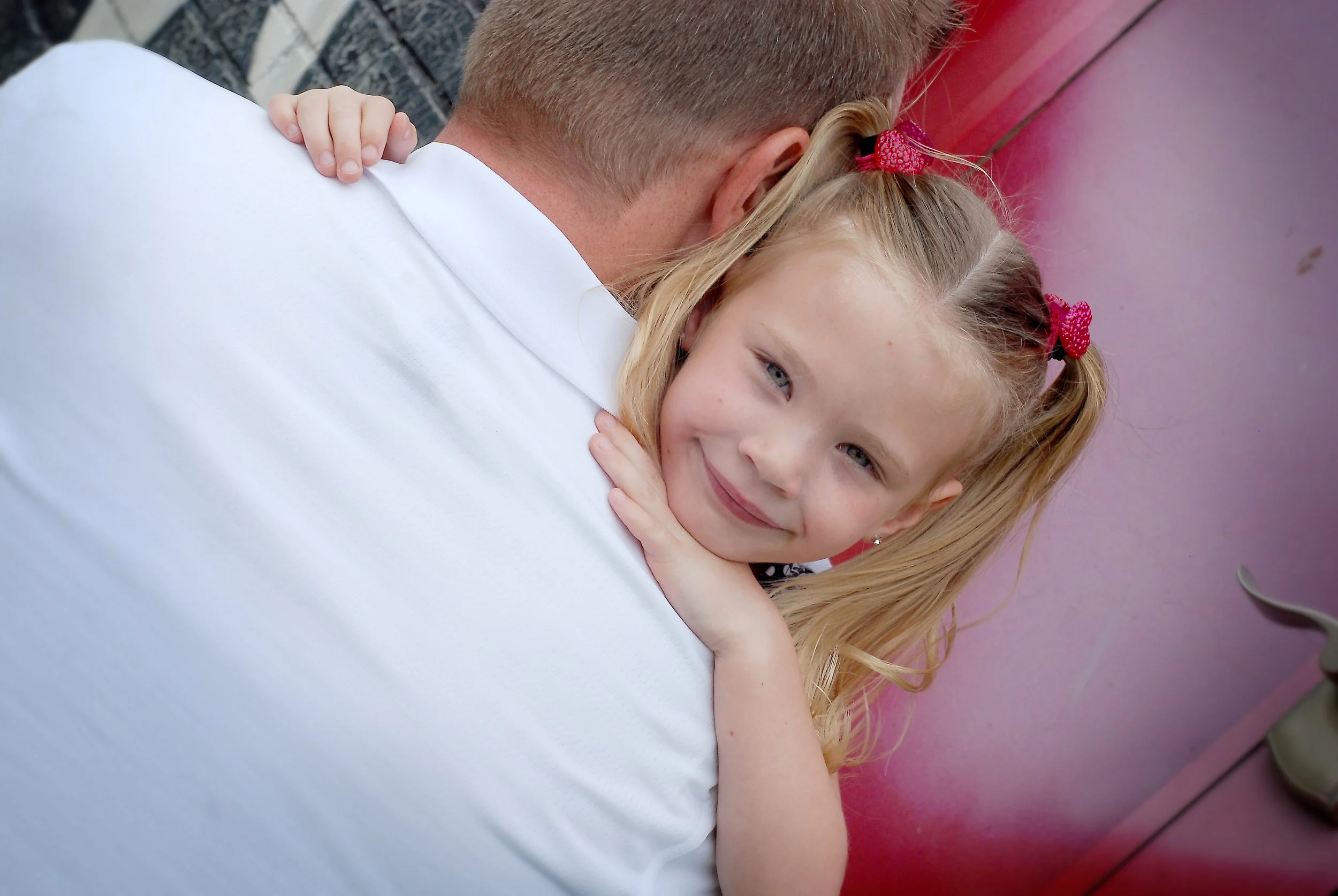 A young girl with blonde hair in two pigtails, smiling and hugging an adult, with her hands resting on the adult's shoulder, against a pink wall.