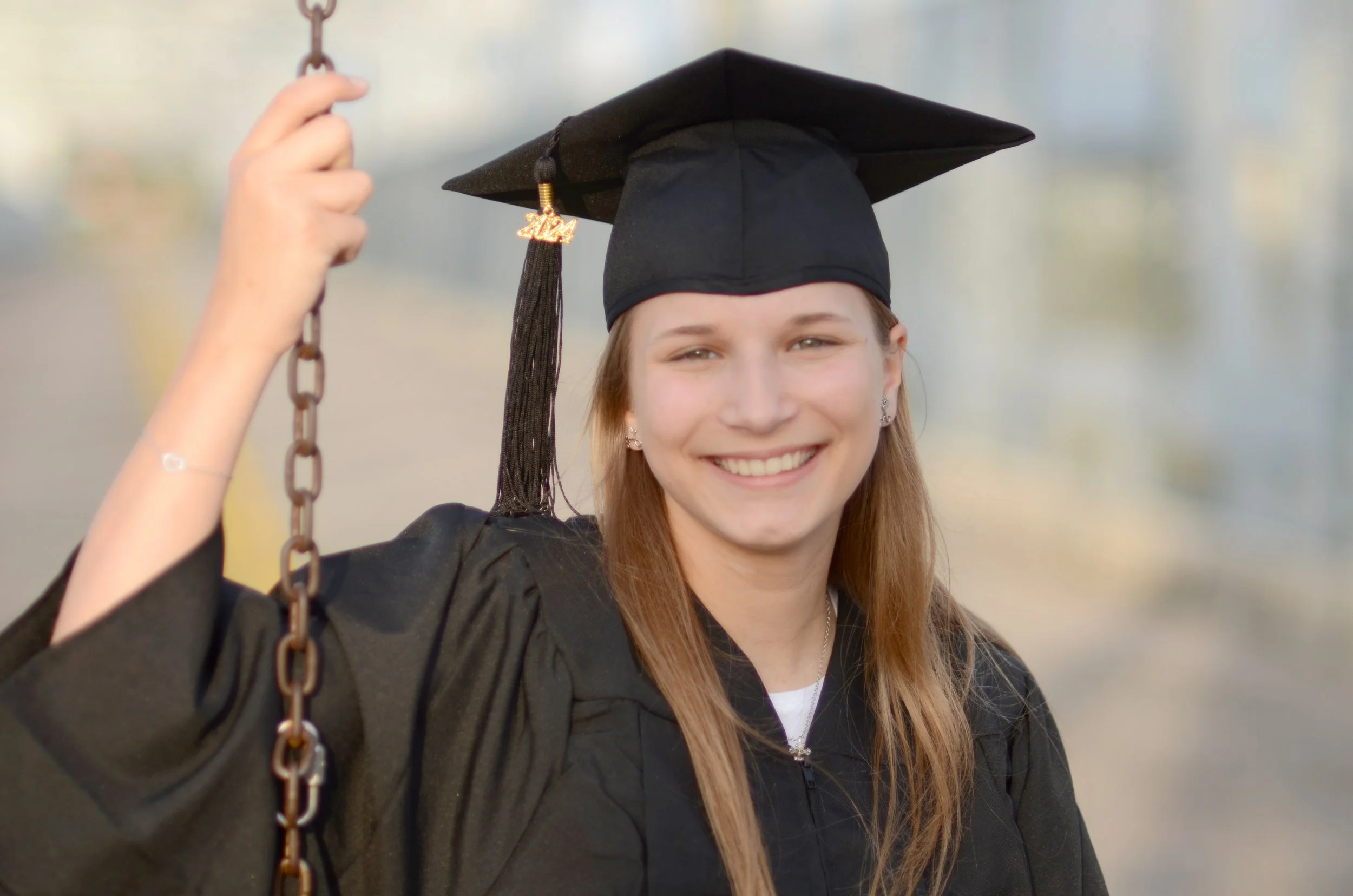 Young woman in a graduation cap and gown smiling, holding a chain, outdoors with blurred background