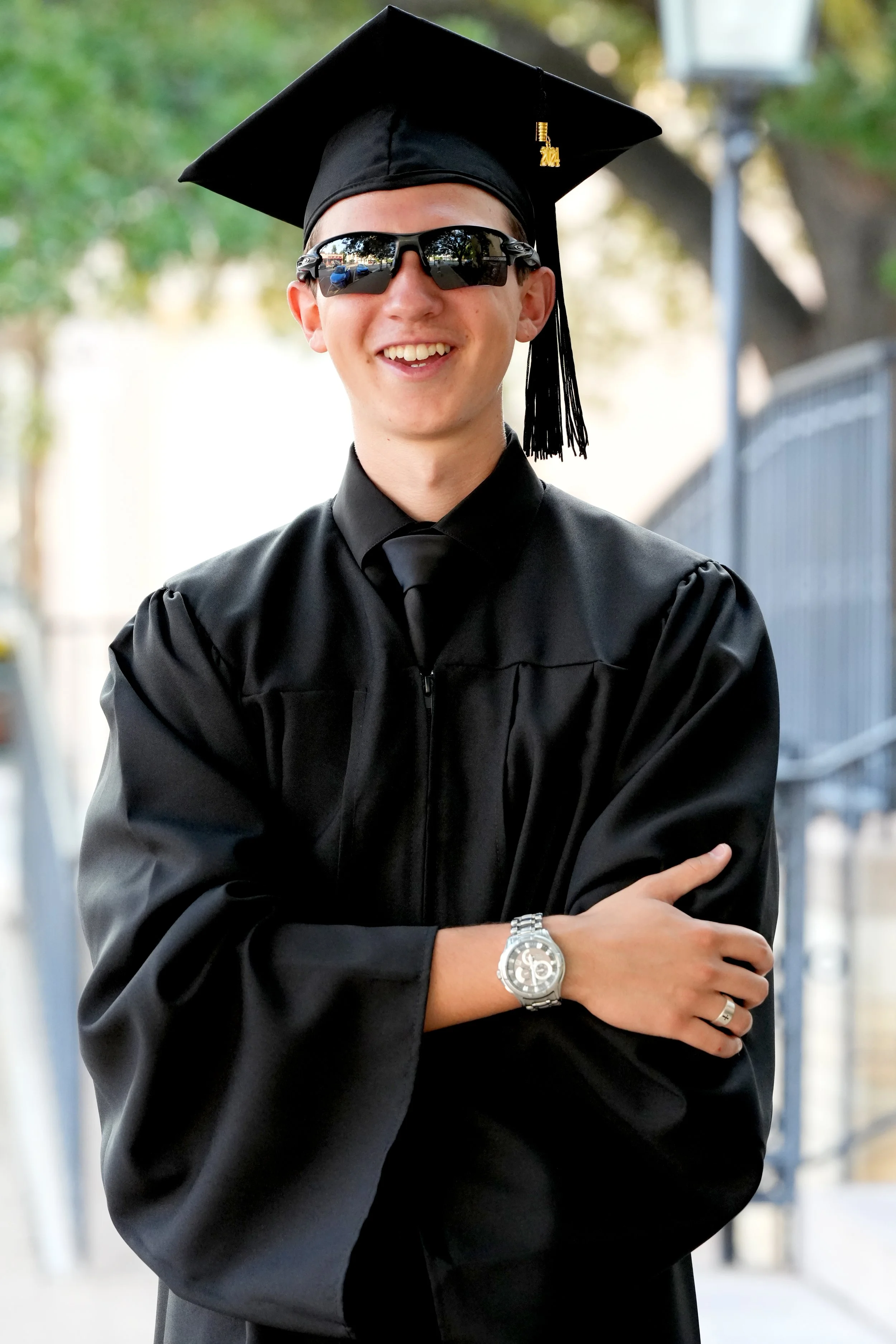 Young man in a graduation cap and gown, wearing sunglasses, smiling outdoors during the daytime.