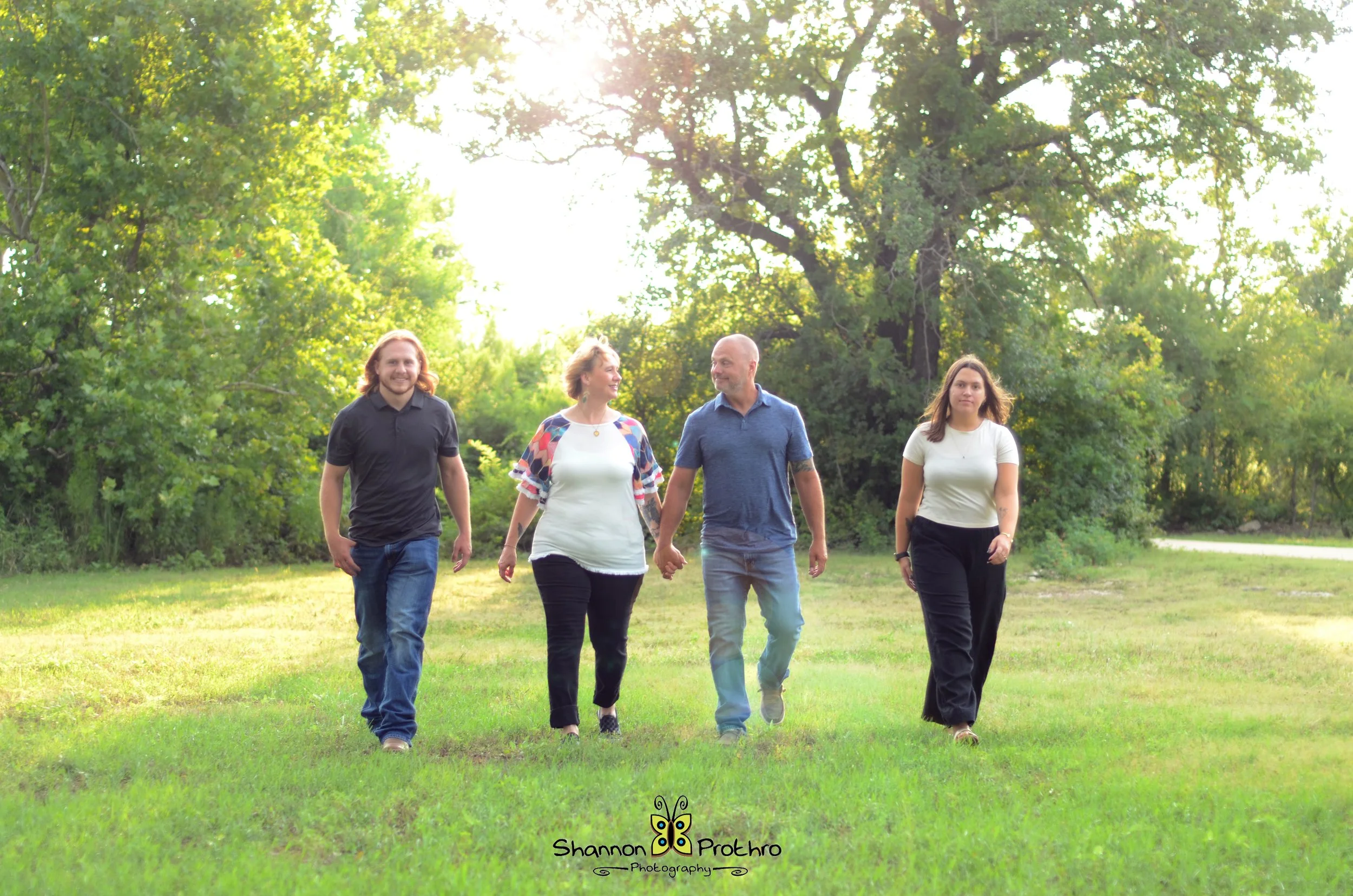 Four people walking hand-in-hand through a green park with trees and sunlight in the background.