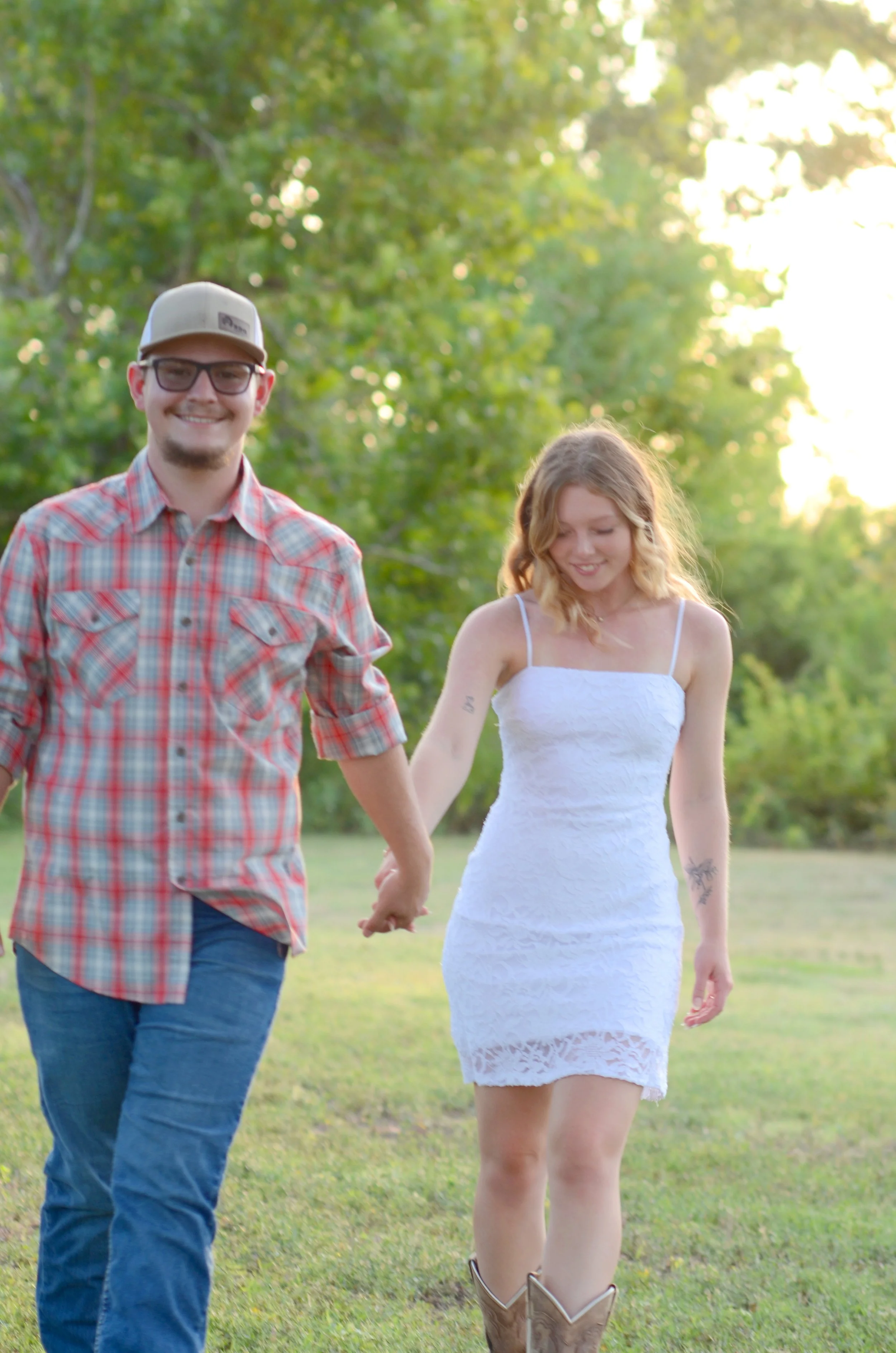 A smiling man in a plaid shirt and baseball cap holding hands with a woman in a white dress walking outdoors in a grassy area with green trees and sunlight in the background.