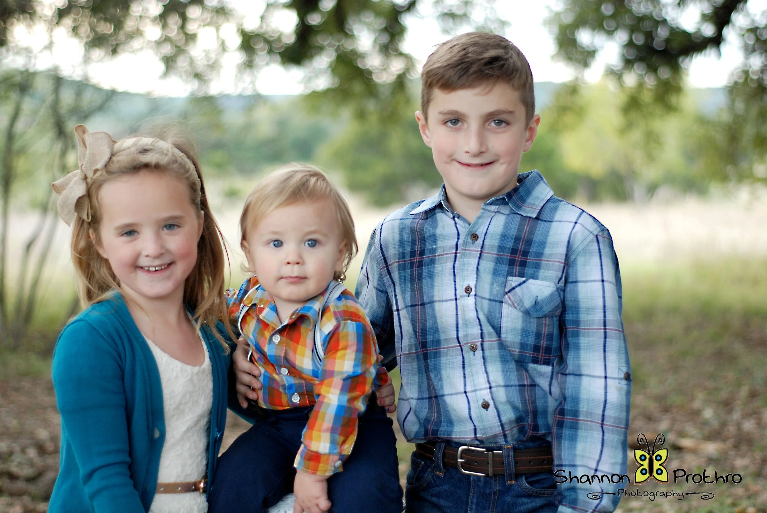 Three children, two girls and one boy, standing outdoors in a park with green trees in the background, smiling at the camera.
