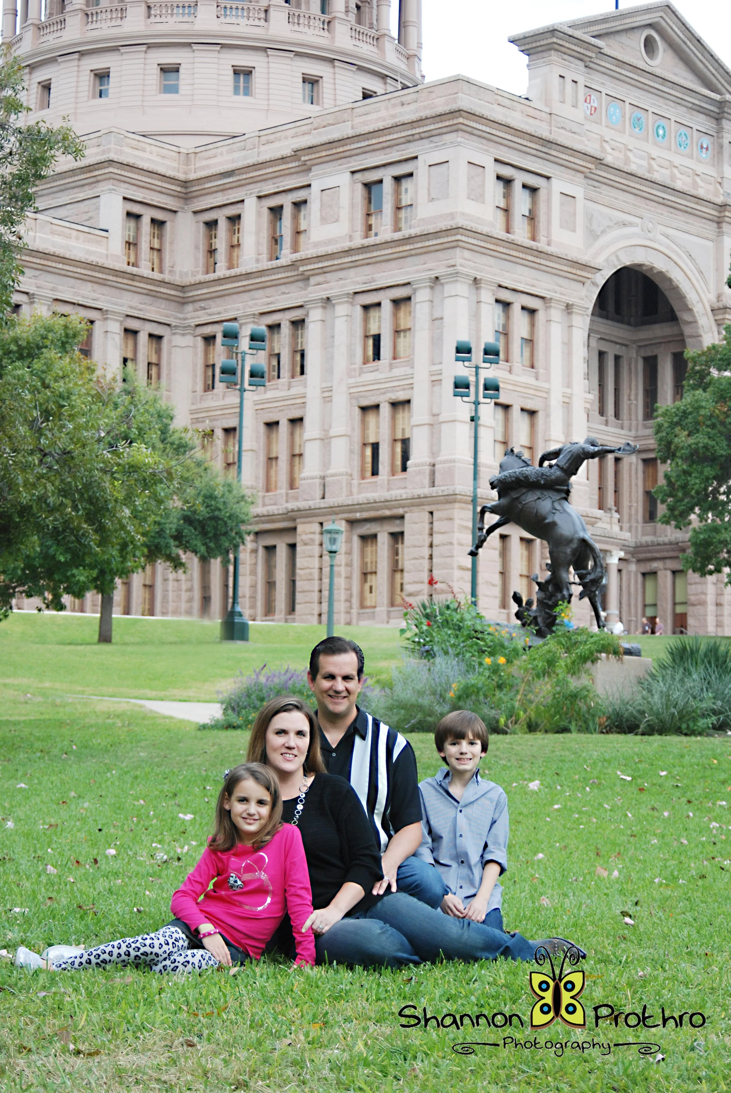 A family of four sitting on the grass in front of the Texas State Capitol building in Austin, Texas.