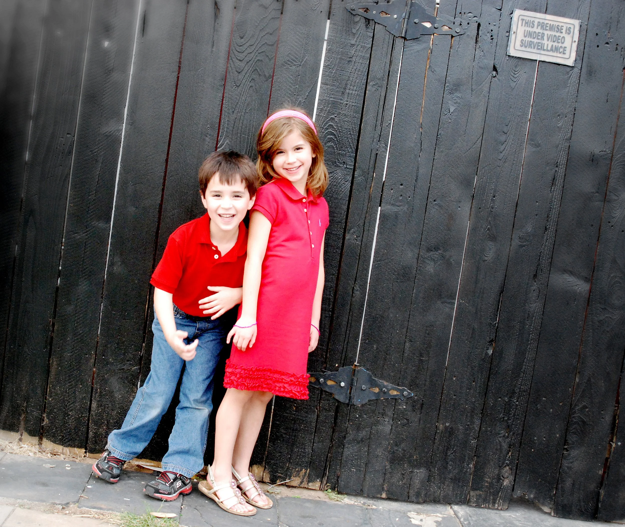 Two children, a boy and a girl, standing in front of a large black wooden gate. The girl is wearing a red dress and sandals, and the boy is wearing a red polo shirt, blue jeans, and sneakers. Both are smiling.