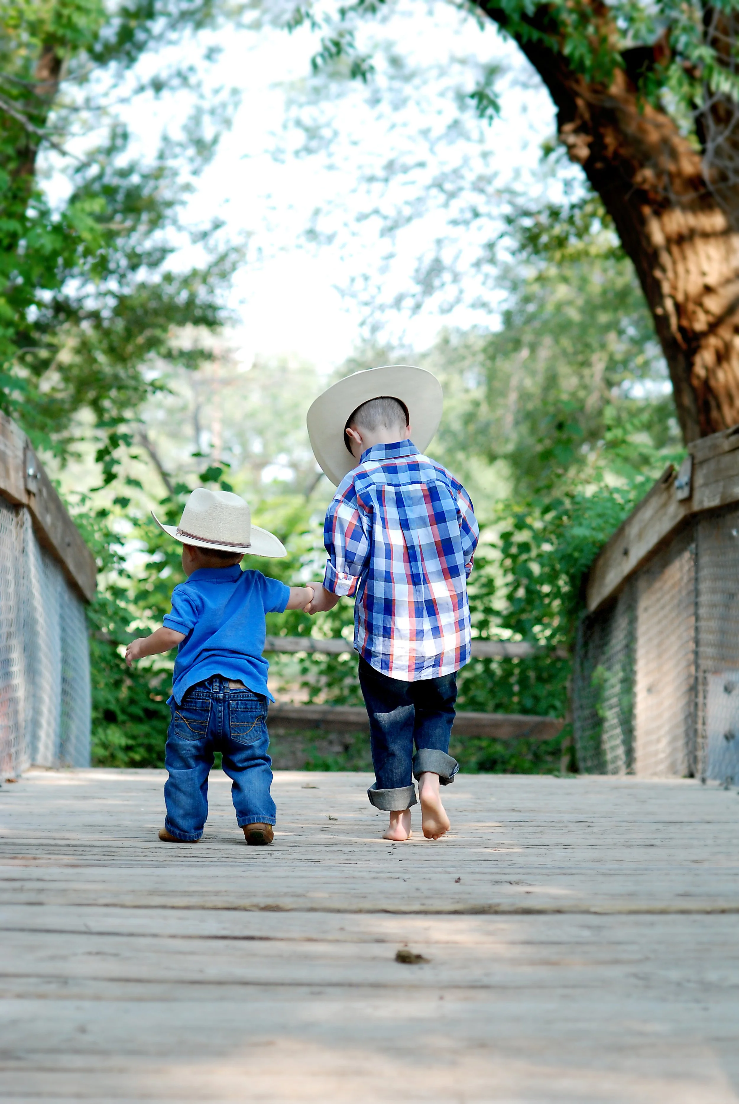 Two children walking hand in hand on a wooden bridge surrounded by trees, both wearing cowboy hats and casual clothes.