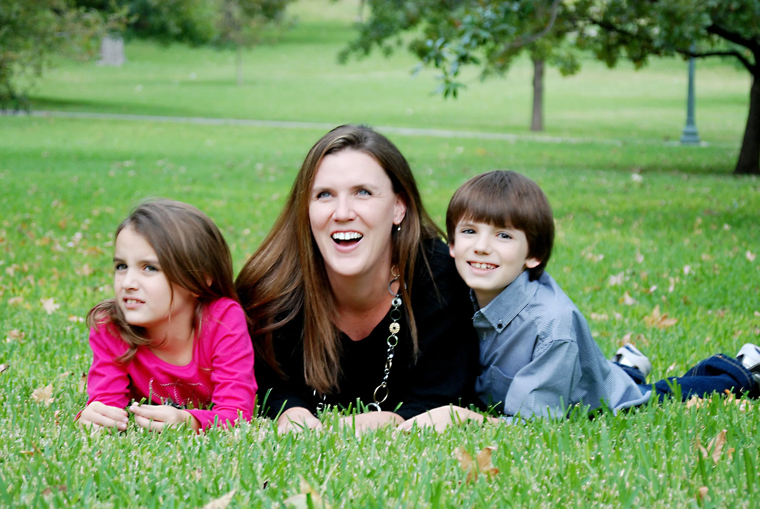 A woman with two young children lying on a grassy park with trees in the background, smiling and enjoying the outdoors.
