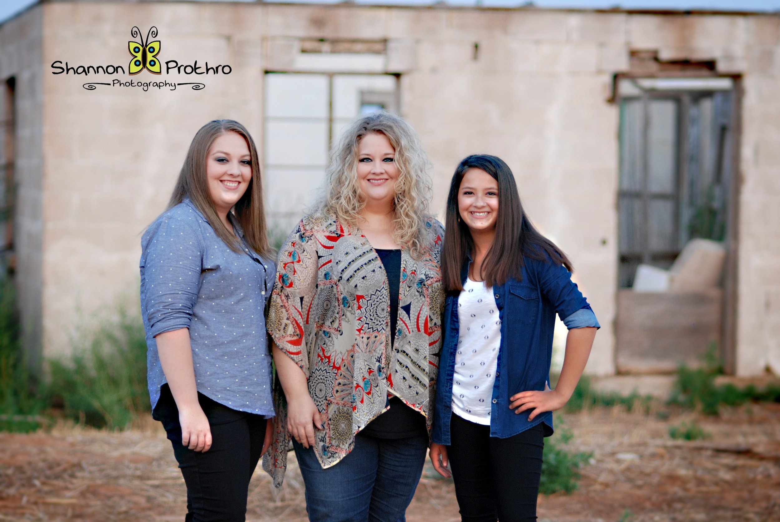 Three women standing outdoors in front of a rustic building with a window and an open doorway, smiling at the camera.