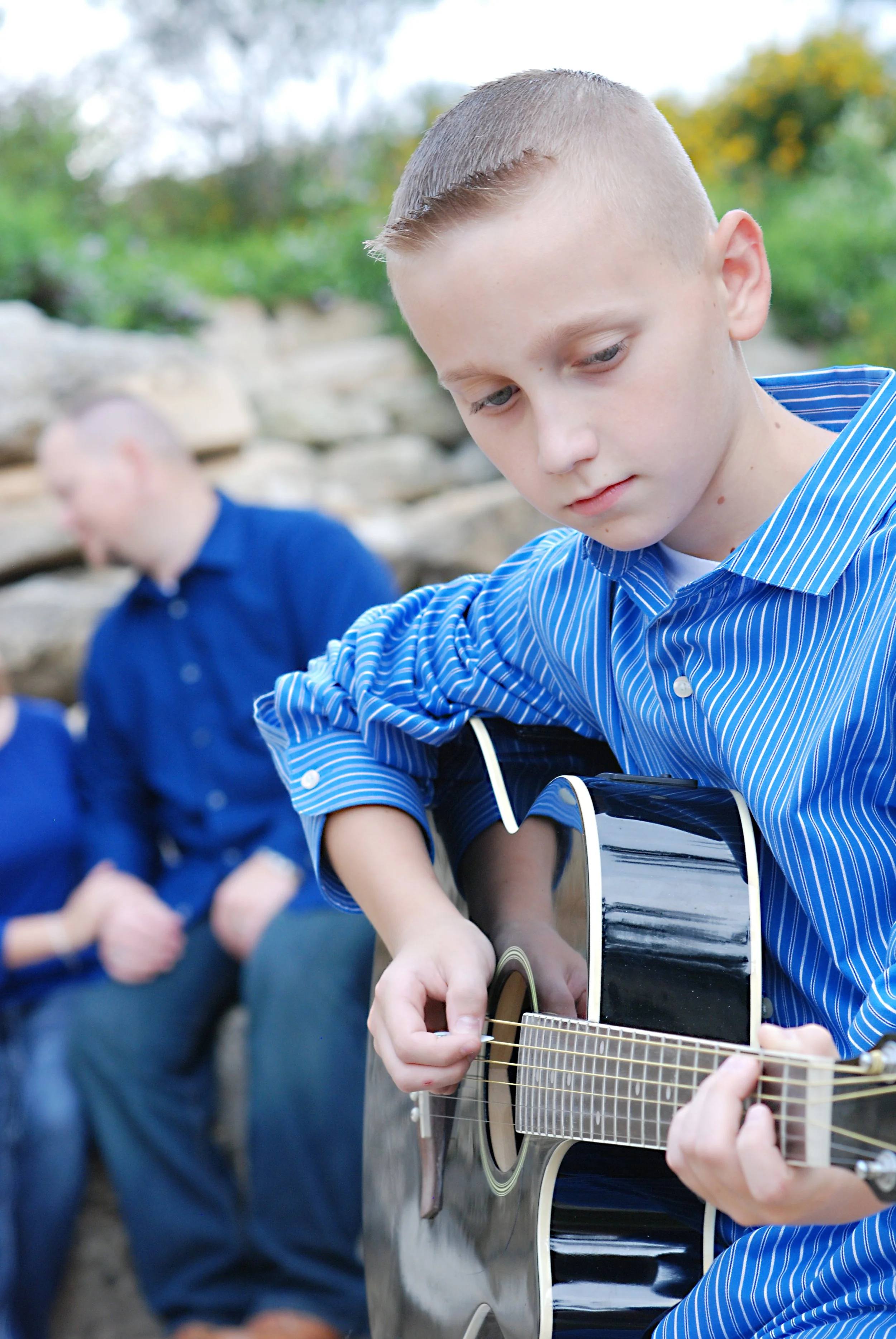 A young boy in a blue striped shirt playing an acoustic guitar outdoors with a group of children in the background.