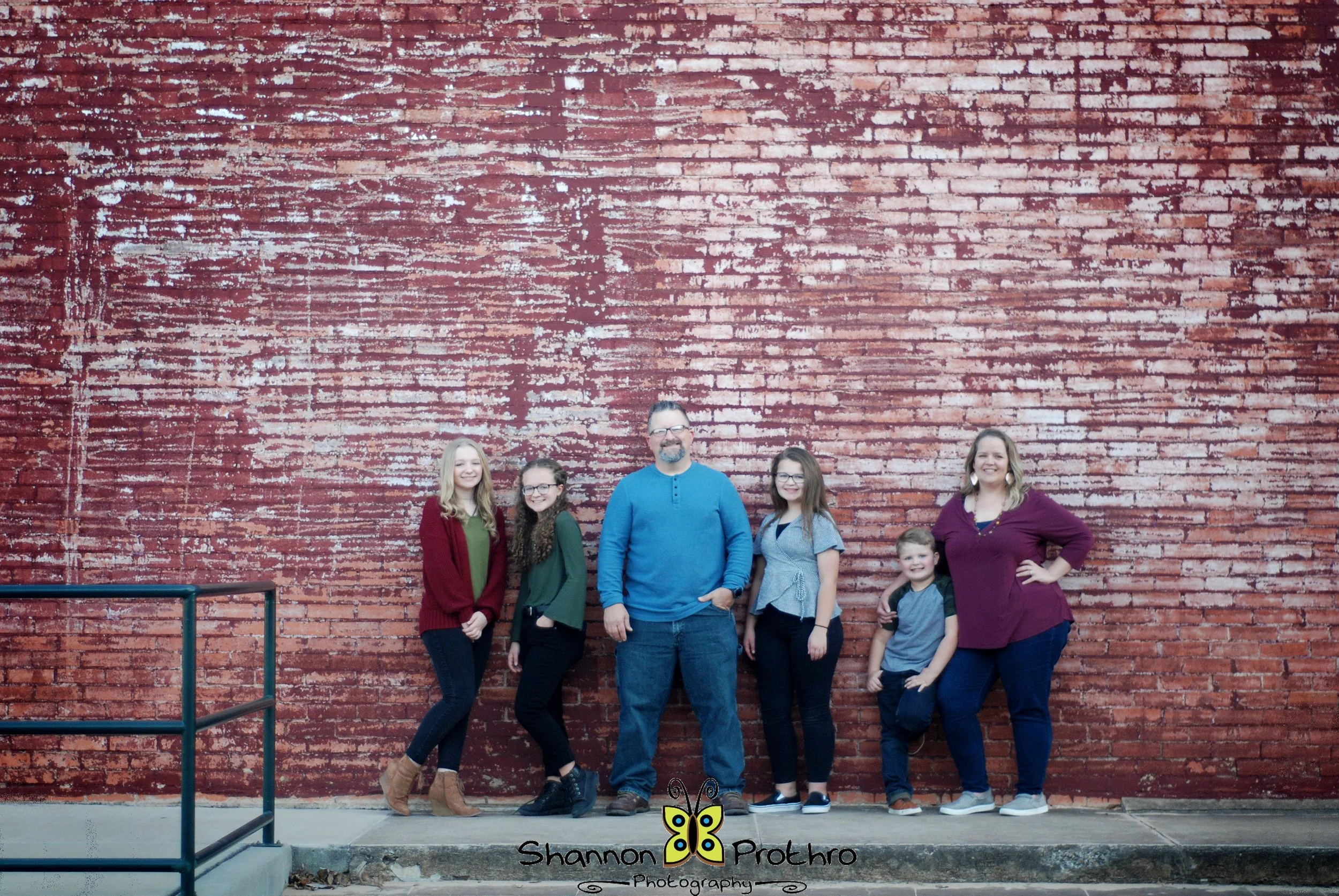 A group of six people, including children and adults, standing in front of a large red brick wall, smiling and posing for a photo.