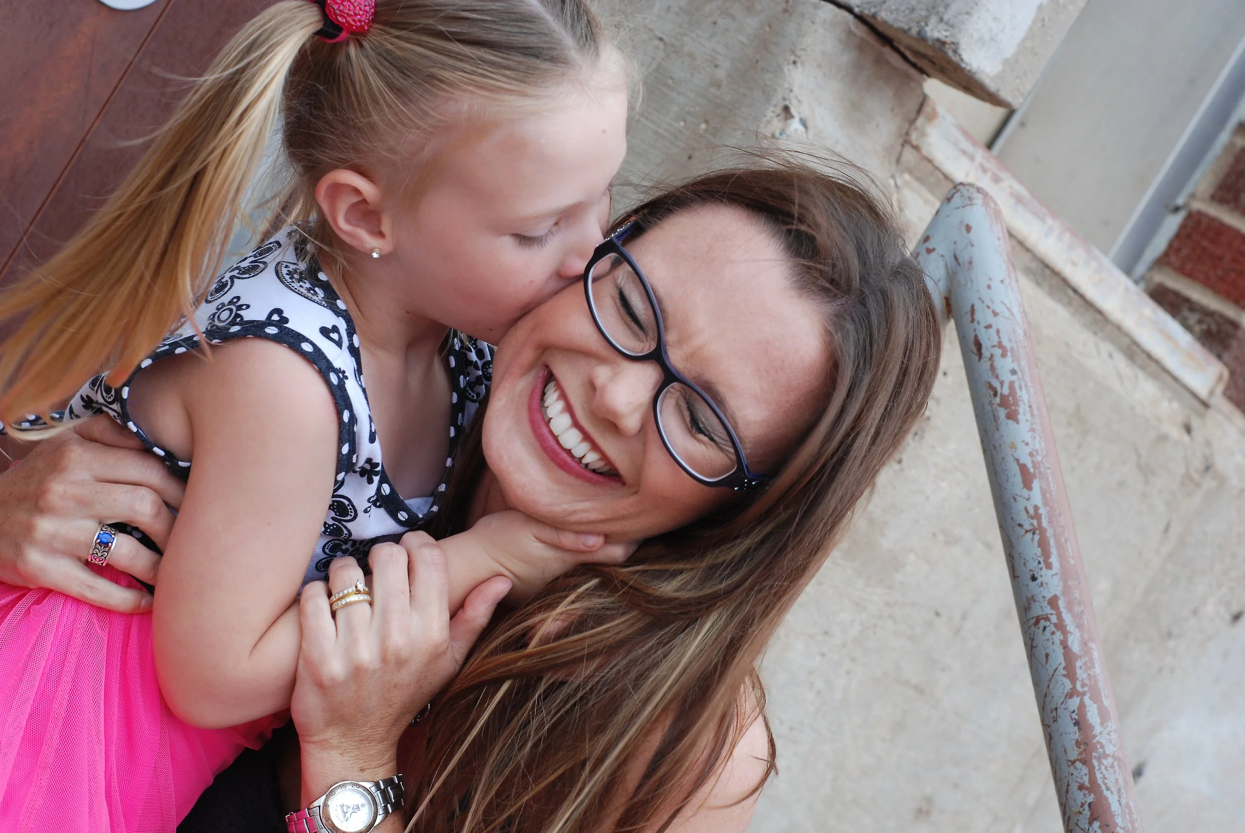 A mother with brown hair and glasses smiling as her young daughter kisses her cheek, outdoors near brick and concrete walls.