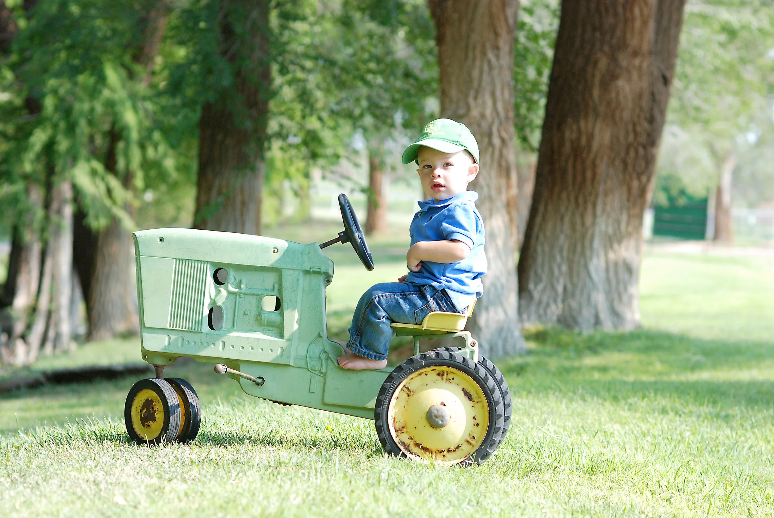 A young boy sitting on a vintage toy tractor outdoors on a grassy area with large trees in the background.