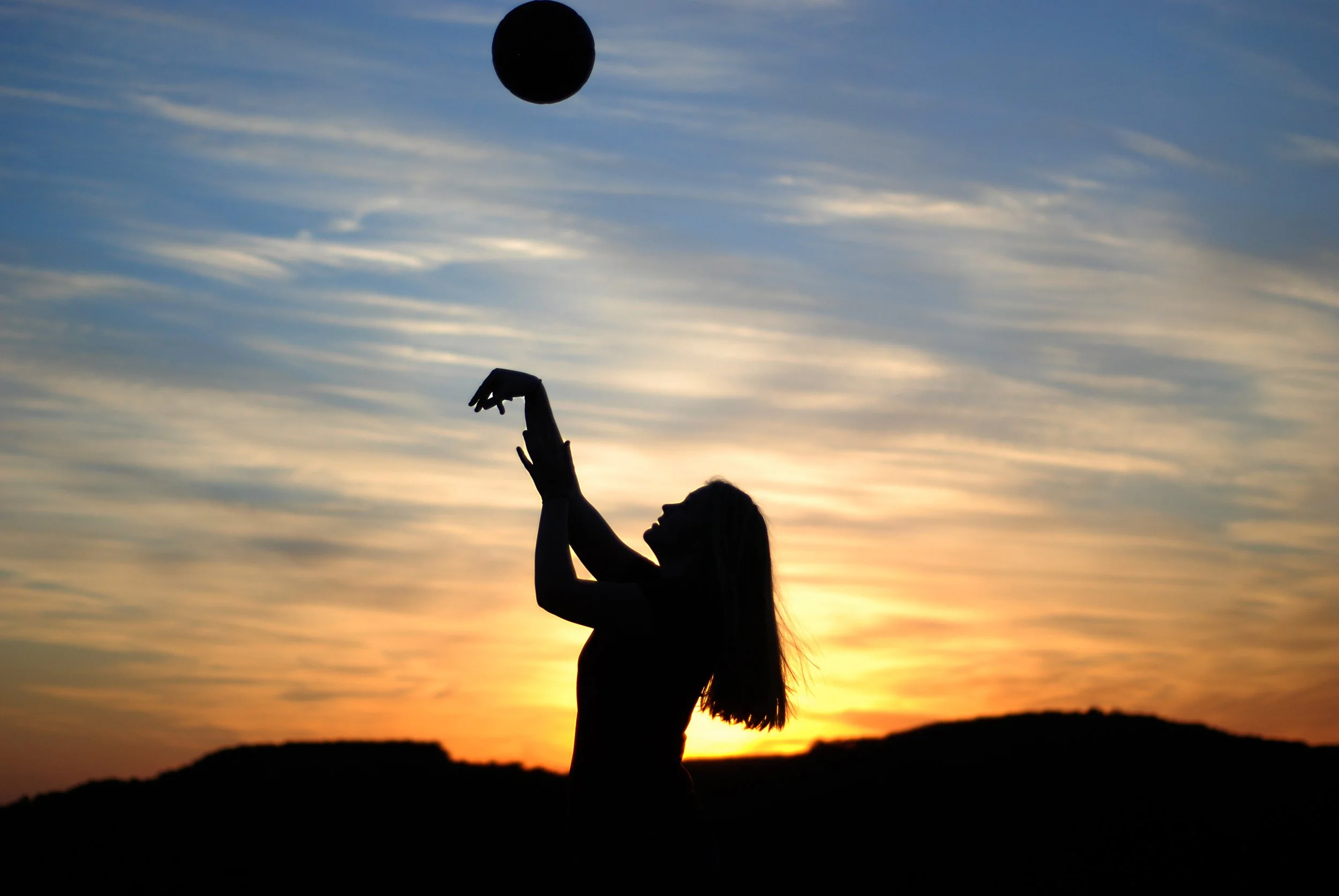 Silhouette of a woman playing with a ball at sunset with a colorful sky and distant hills in the background.