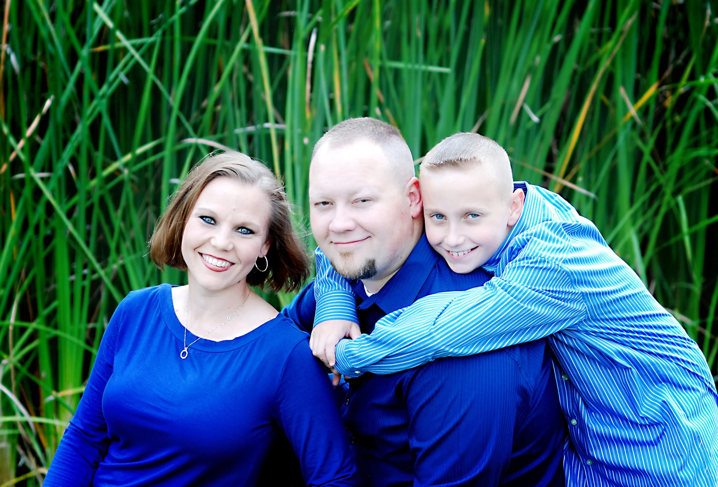 A woman and two boys wearing blue shirts smiling, with tall green grass in the background.