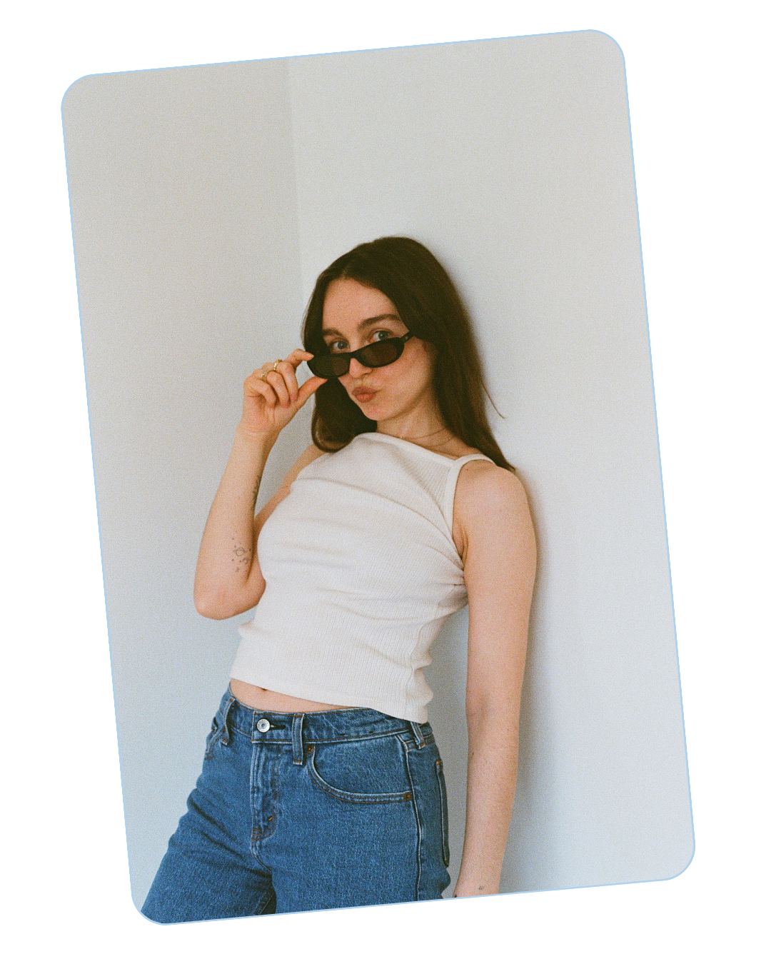 Girl with brunette hair wearing a white tank top, blue jeans, and sunglasses making a face at the camera.