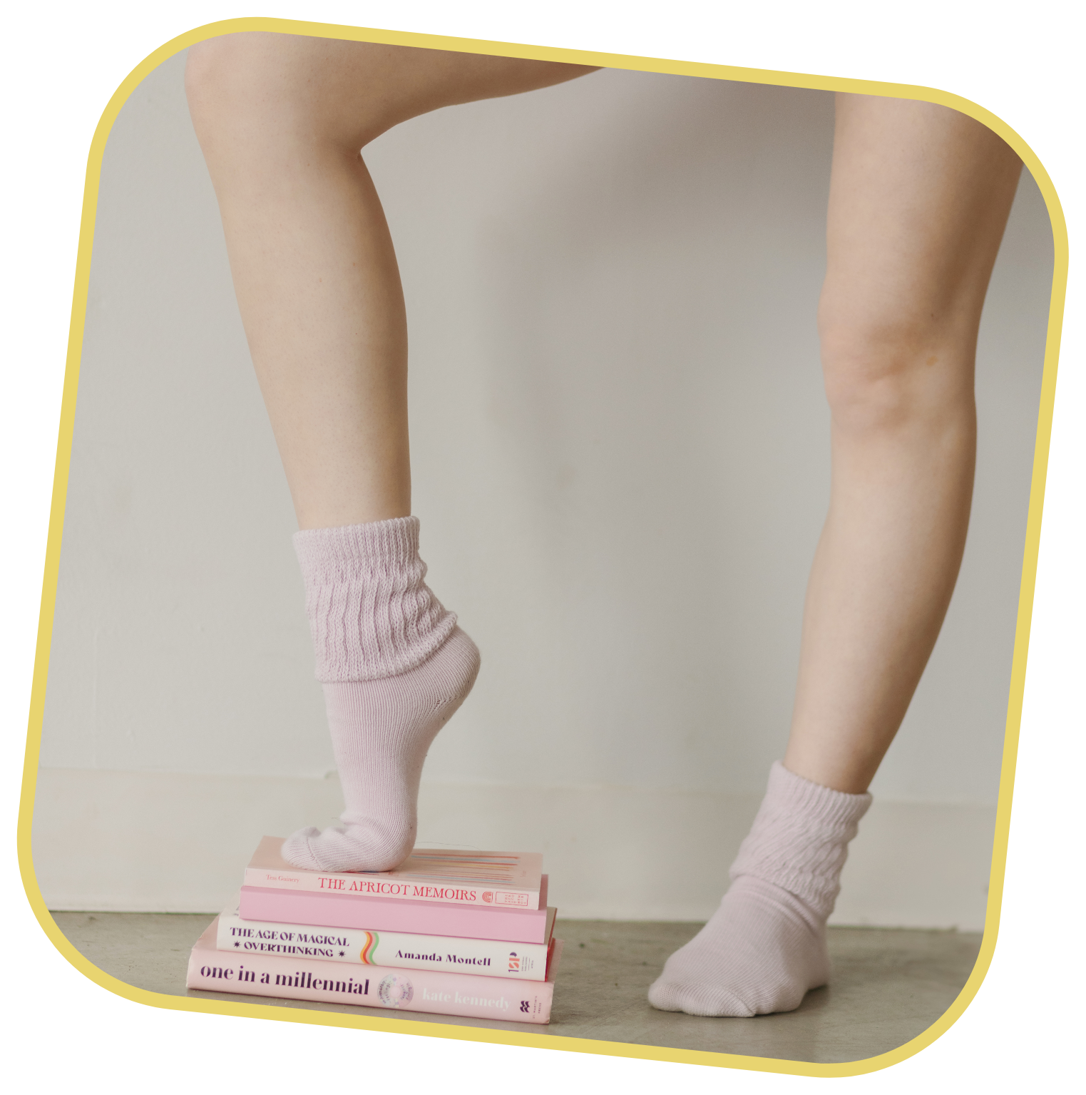 Taylor standing on a stack of books, wearing pink socks. One foot is on the top book, the other on the ground.
