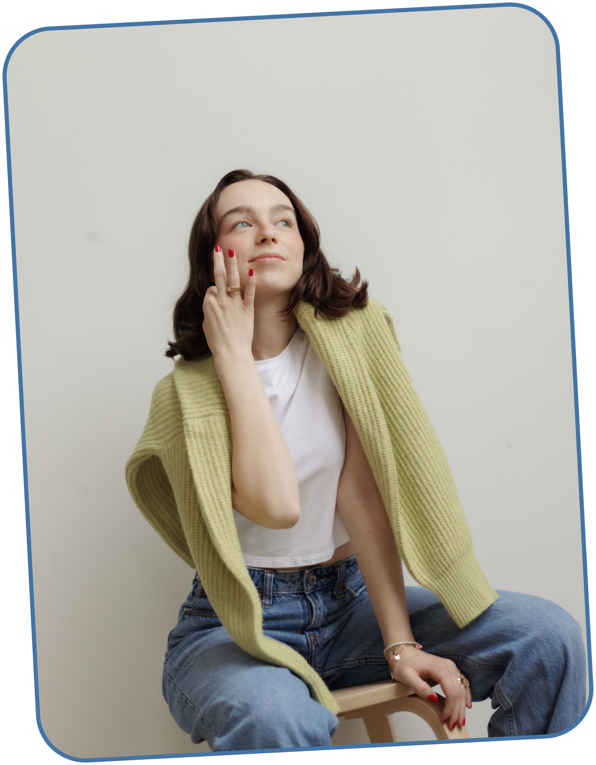 Taylor, the copywriter, wears a white t-shirt, blue jeans, and a green cardigan draped over her shoulders, and sits on a wooden stool against a plain white background.