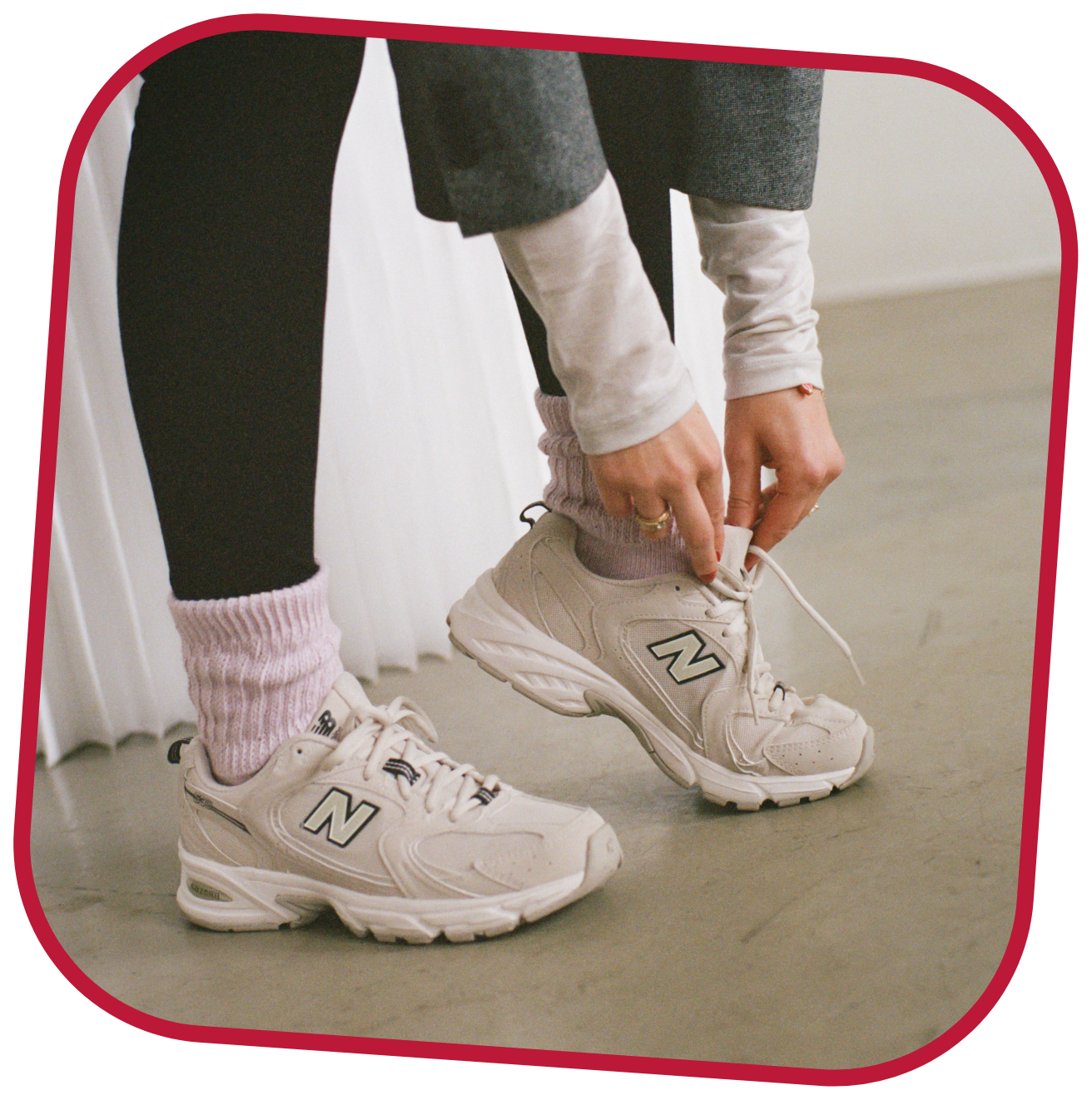 Person tying their beige New Balance sneakers, wearing pink socks, black leggings, white top and a grey blazer.