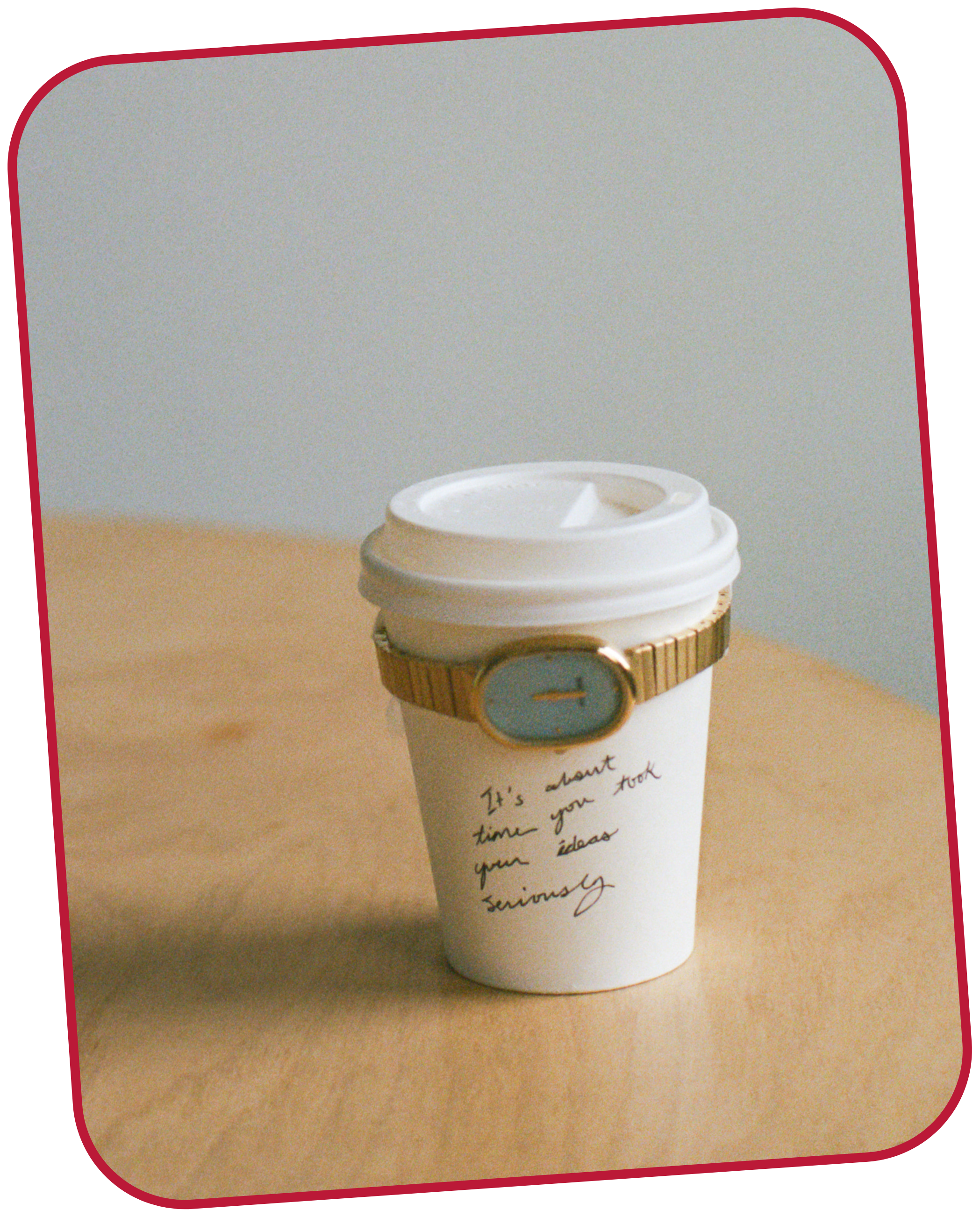 A white to-go coffee cup with handwritten text that says, "It's about time you take your ideas seriously" on a light wooden table and a gold watch with a sky blue face wrapped around the cup.