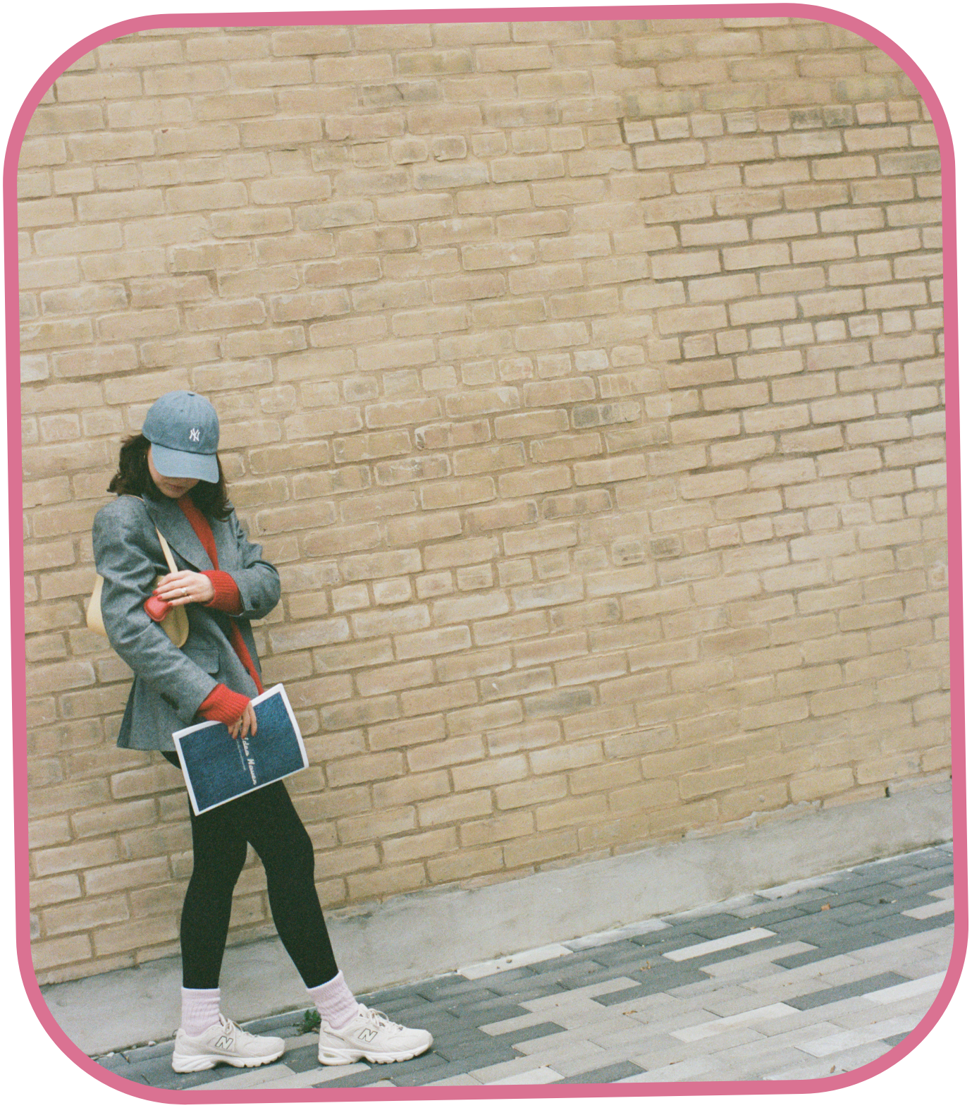 Taylor, The Idea Haven’s copywriter, standing against a brick wall, wearing a blue baseball cap, grey blazer, red sweater, black leggings, white sneakers, and holding a magazine and beige purse.