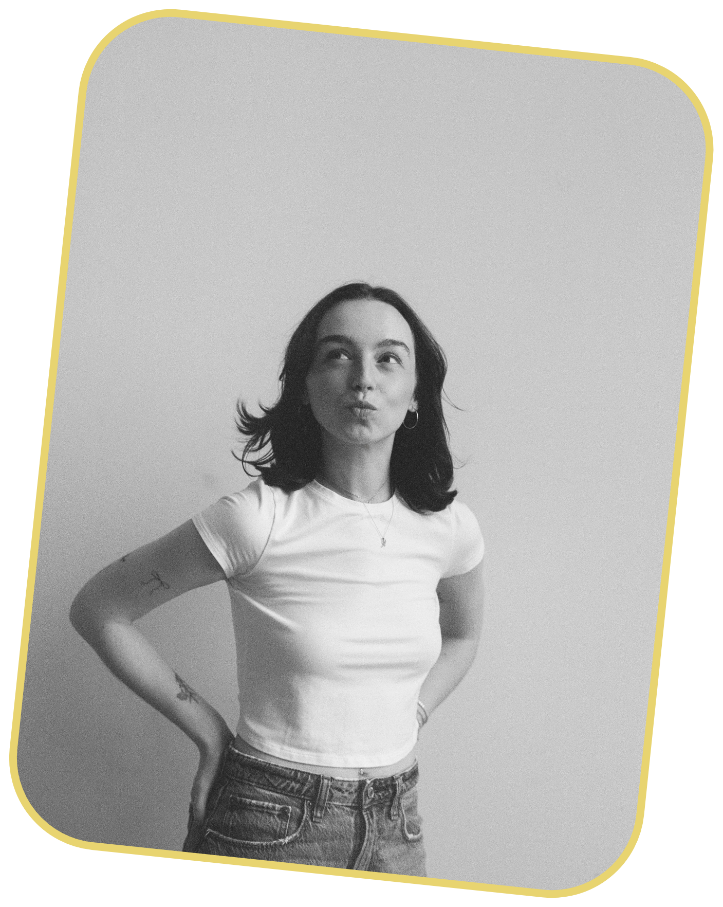 Black and white photo of Taylor with shoulder-length dark hair, wearing a white t-shirt and jeans, standing against a plain wall, with hands on her hip and making a playful, puckered lip expression.