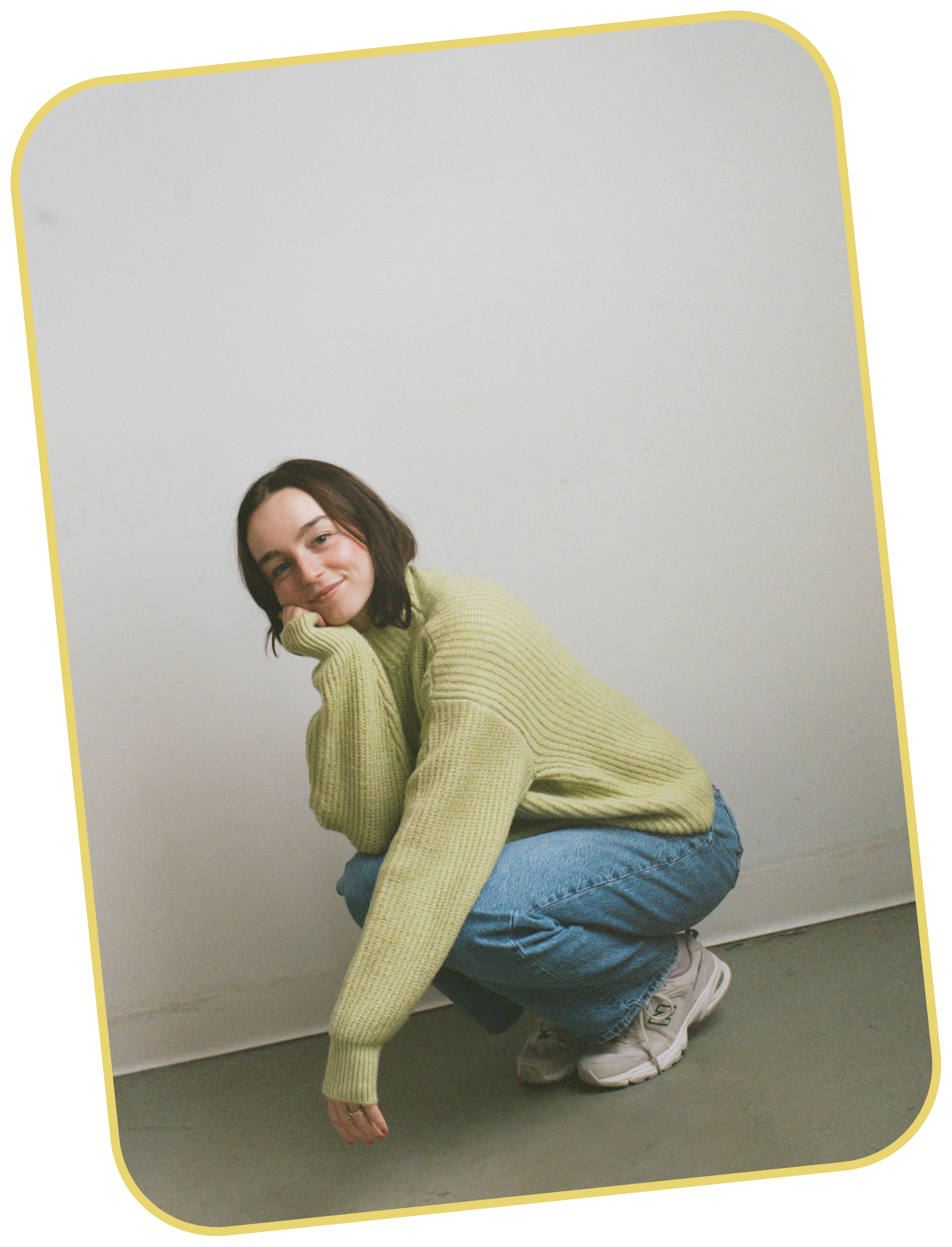 Girl wearing a green sweater, blue jeans, and sneakers squatting down with right elbow leaning on her right knee and her left arm resting against her left knee.