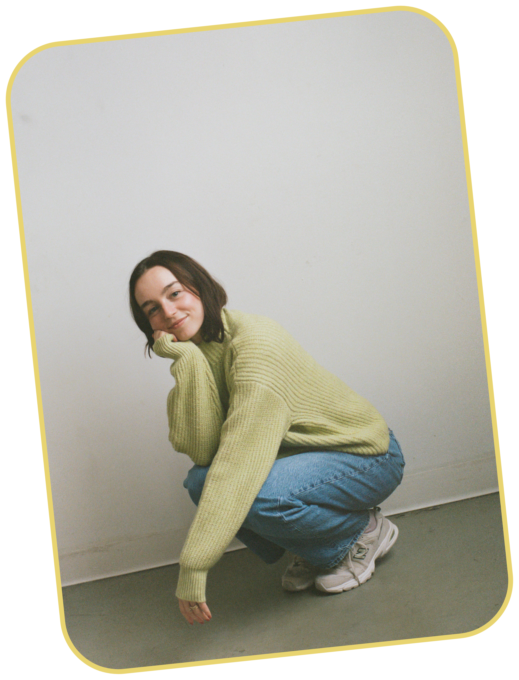 Girl in green sweater, blue jeans, and sneakers crouched down smiling at the camera. One arm is rested on her knee with her hand to her chin and the other arm is loosely draped over knee.