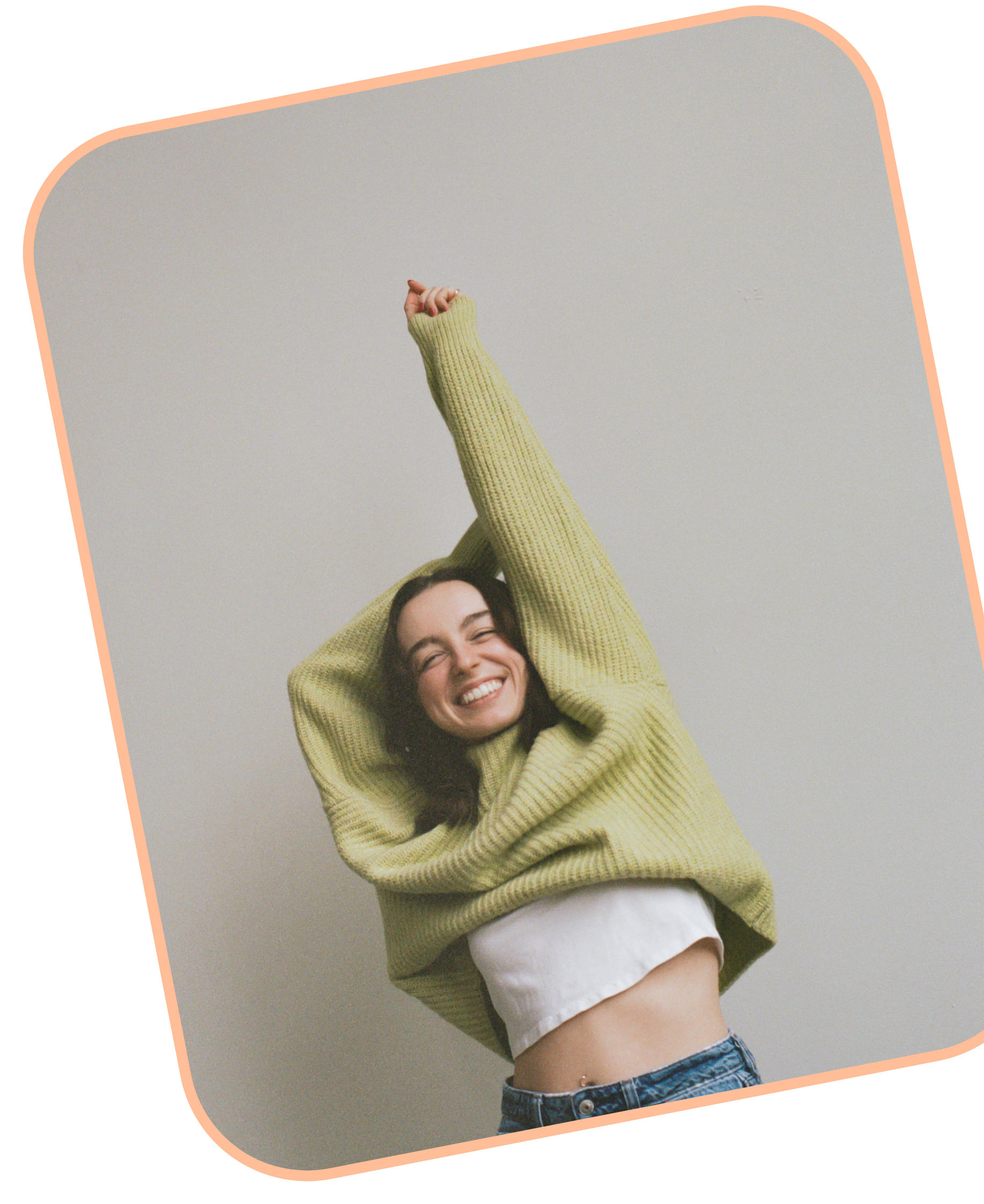 Taylor smiling and wearing a light green knit sweater, white shirt, and blue jeans holding her left arm above her head and the other bent behind her head.