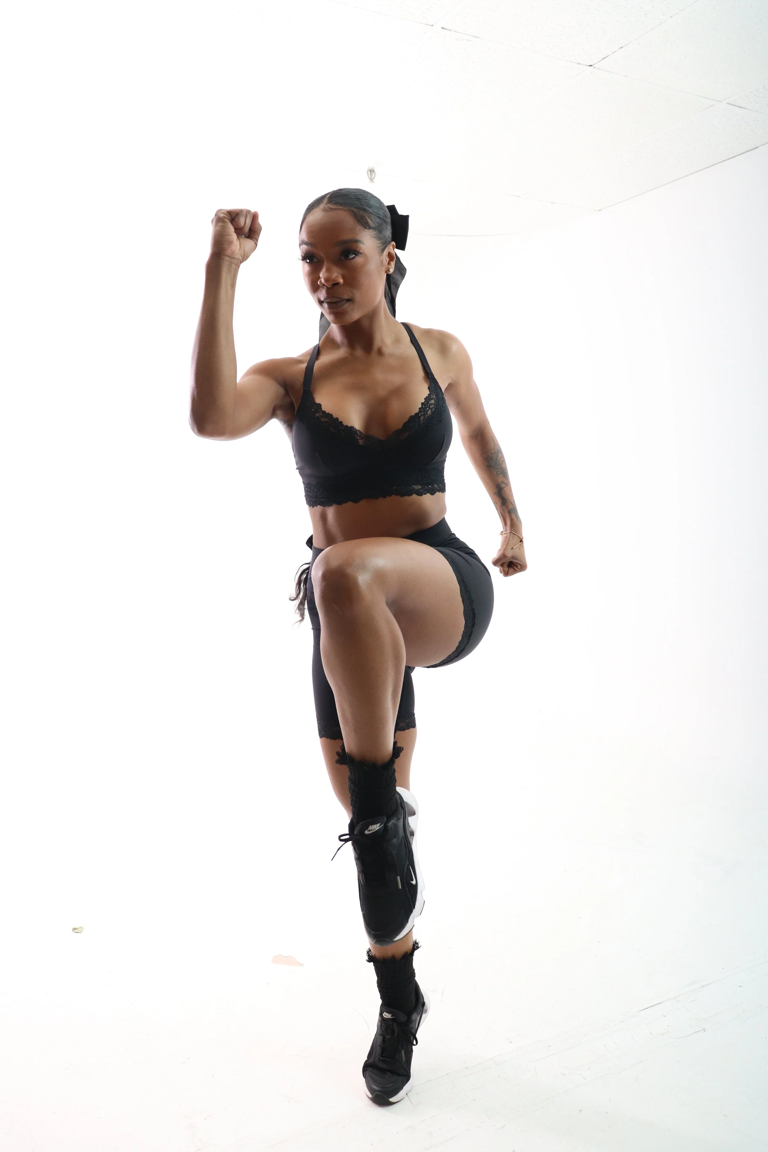 A woman in black athletic wear striking a pose with one leg raised and her arm flexed, against a plain white background.