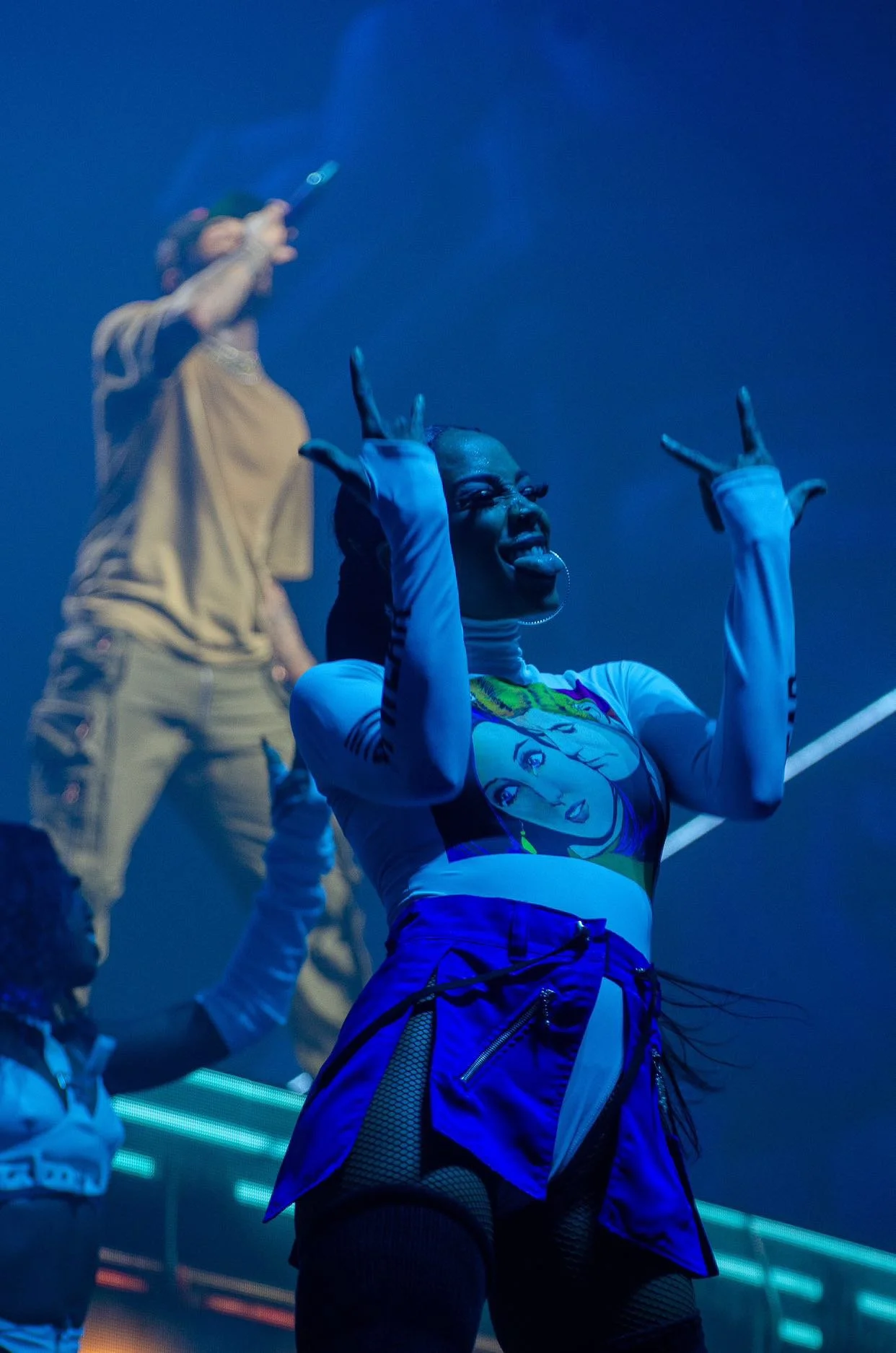 A woman with dark skin making a playful face and gestures in front of a blue background, wearing a comic book style shirt and a blue skirt, with people in the background taking pictures.